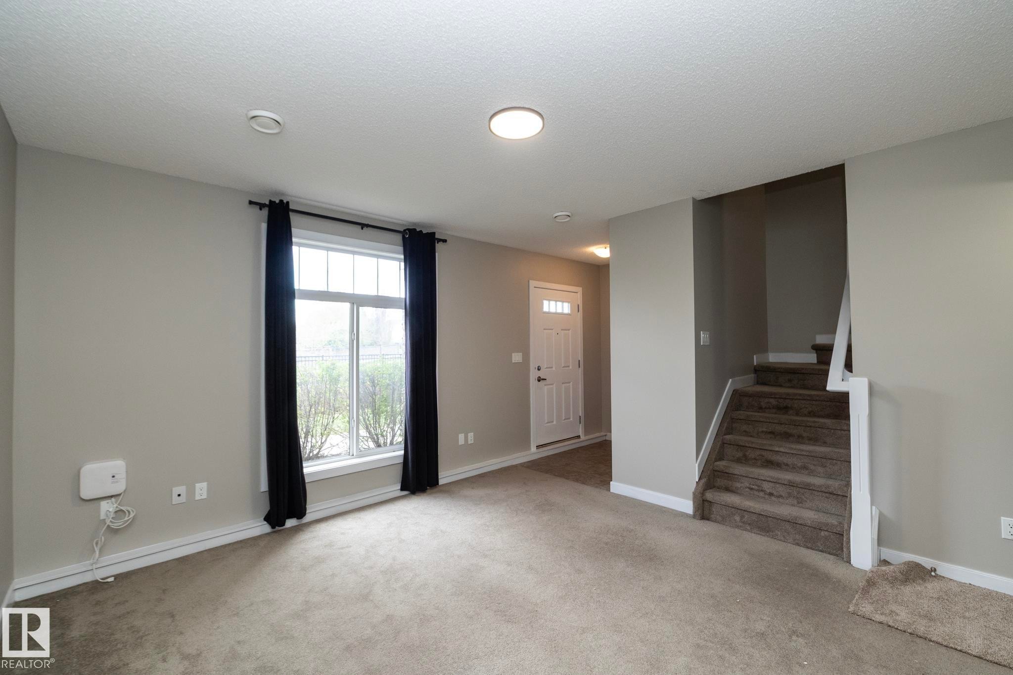 Open concept living space featuring neutral gray walls, plush carpet flooring, a multi-pane window with black drapery, and a visible staircase with carpeting - 117 Callaghan Drive Sw, Edmonton, AB - Indoor Photo Showing Other Room