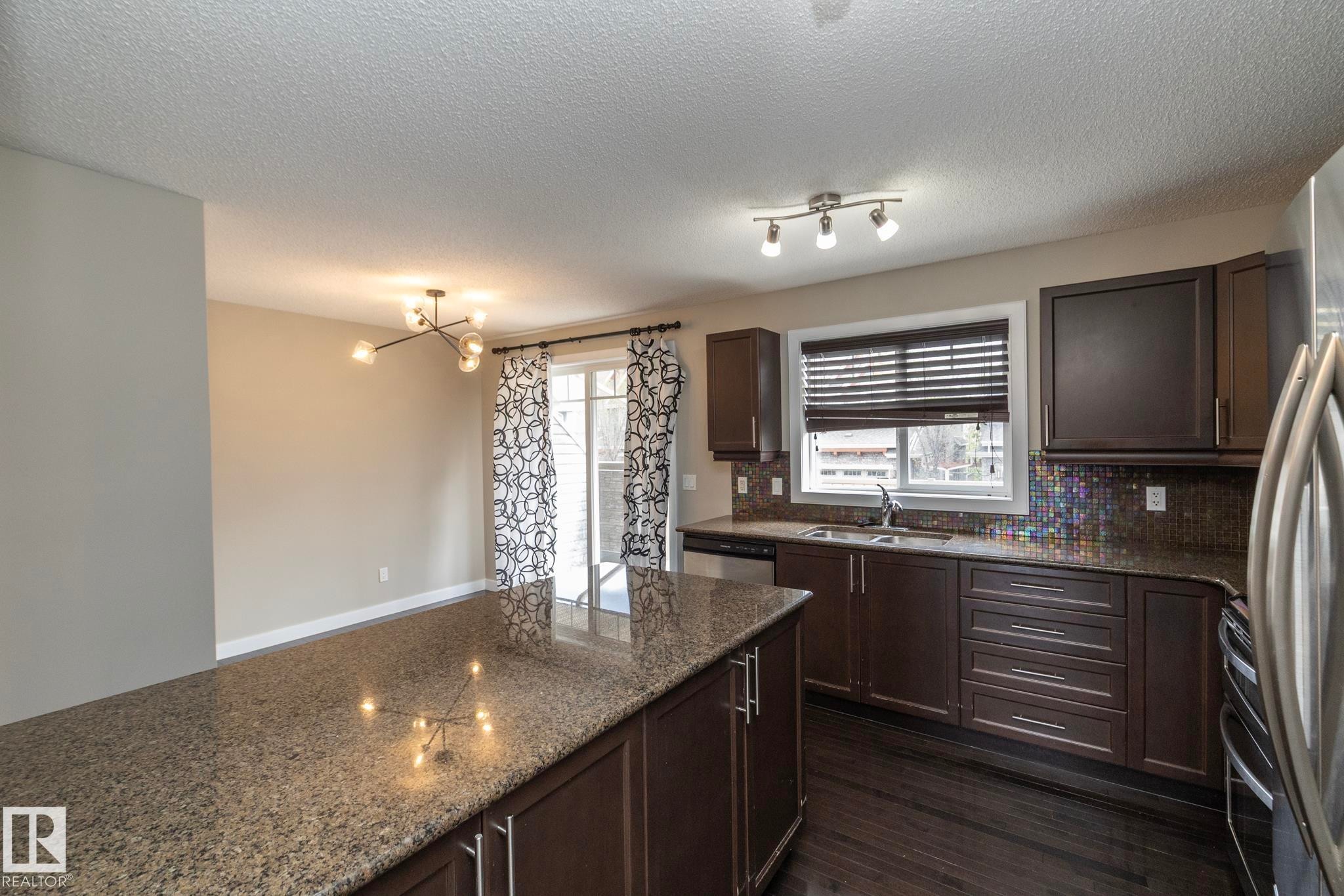 Kitchen featuring an expansive island with a granite countertop, dark wood-finish cabinetry, stainless steel appliances, a mosaic tile backsplash, and dark wood-finish flooring - 117 Callaghan Drive Sw, Edmonton, AB - Indoor Photo Showing Kitchen With Double Sink
