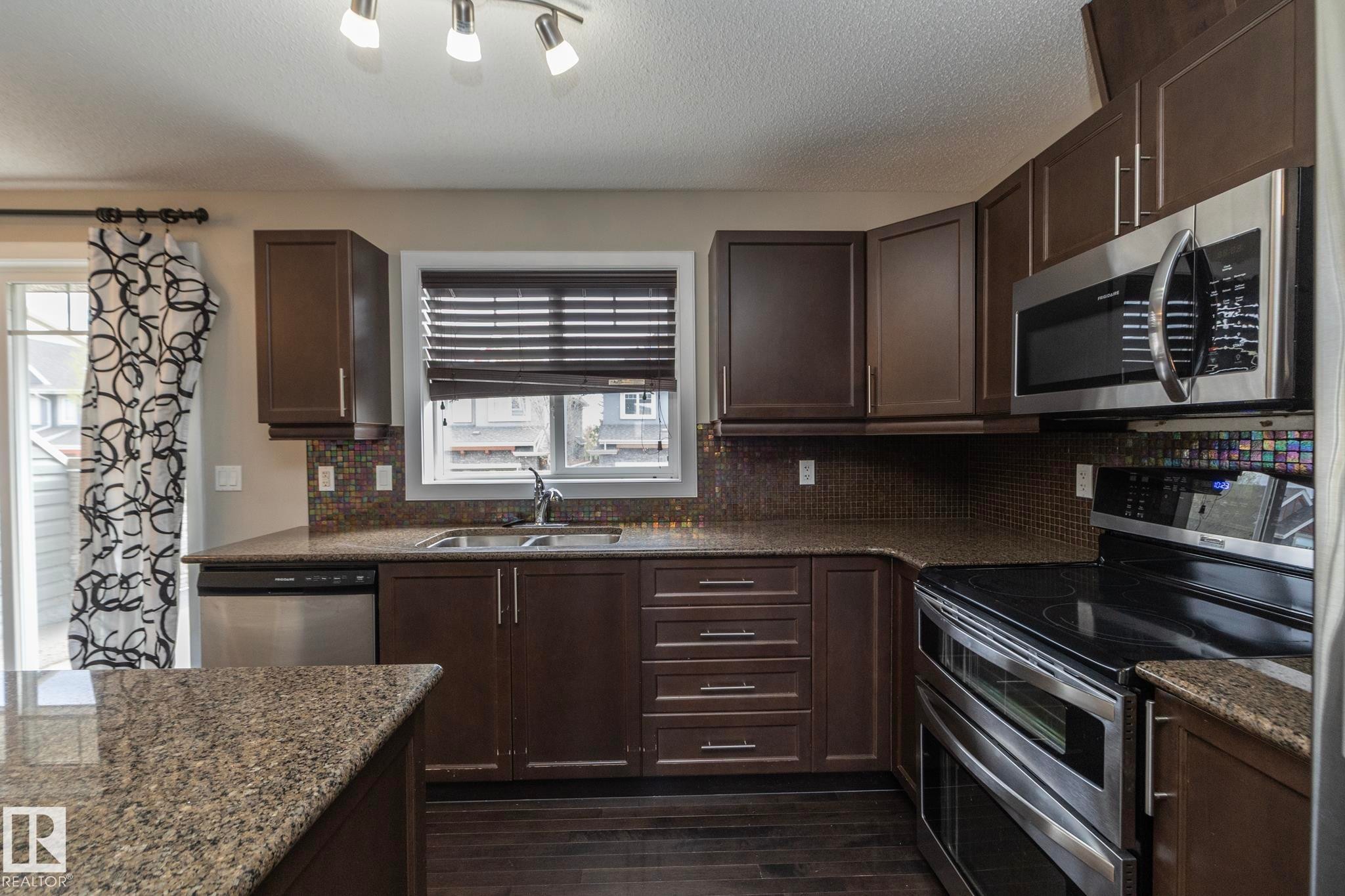 Contemporary kitchen featuring dark wood cabinetry, granite-finish countertops, stainless steel appliances, a mosaic tile backsplash, and dark wood-finish flooring - 117 Callaghan Drive Sw, Edmonton, AB - Indoor Photo Showing Kitchen With Double Sink