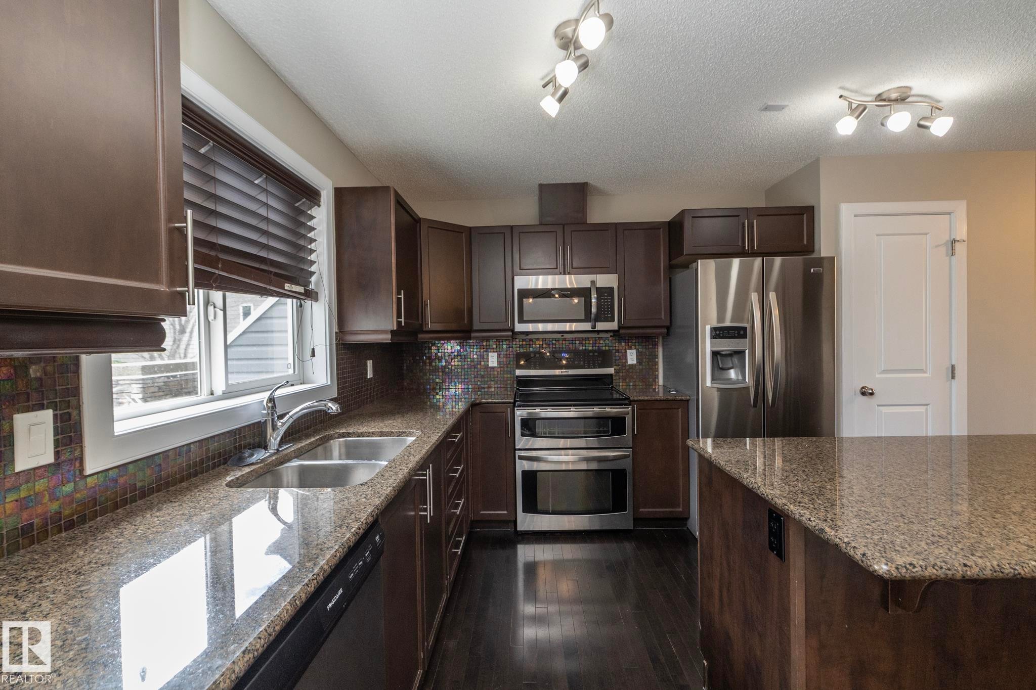 Kitchen featuring dark wood cabinetry, stainless steel appliances, granite countertops, and a mosaic tile backsplash - 117 Callaghan Drive Sw, Edmonton, AB - Indoor Photo Showing Kitchen With Double Sink With Upgraded Kitchen
