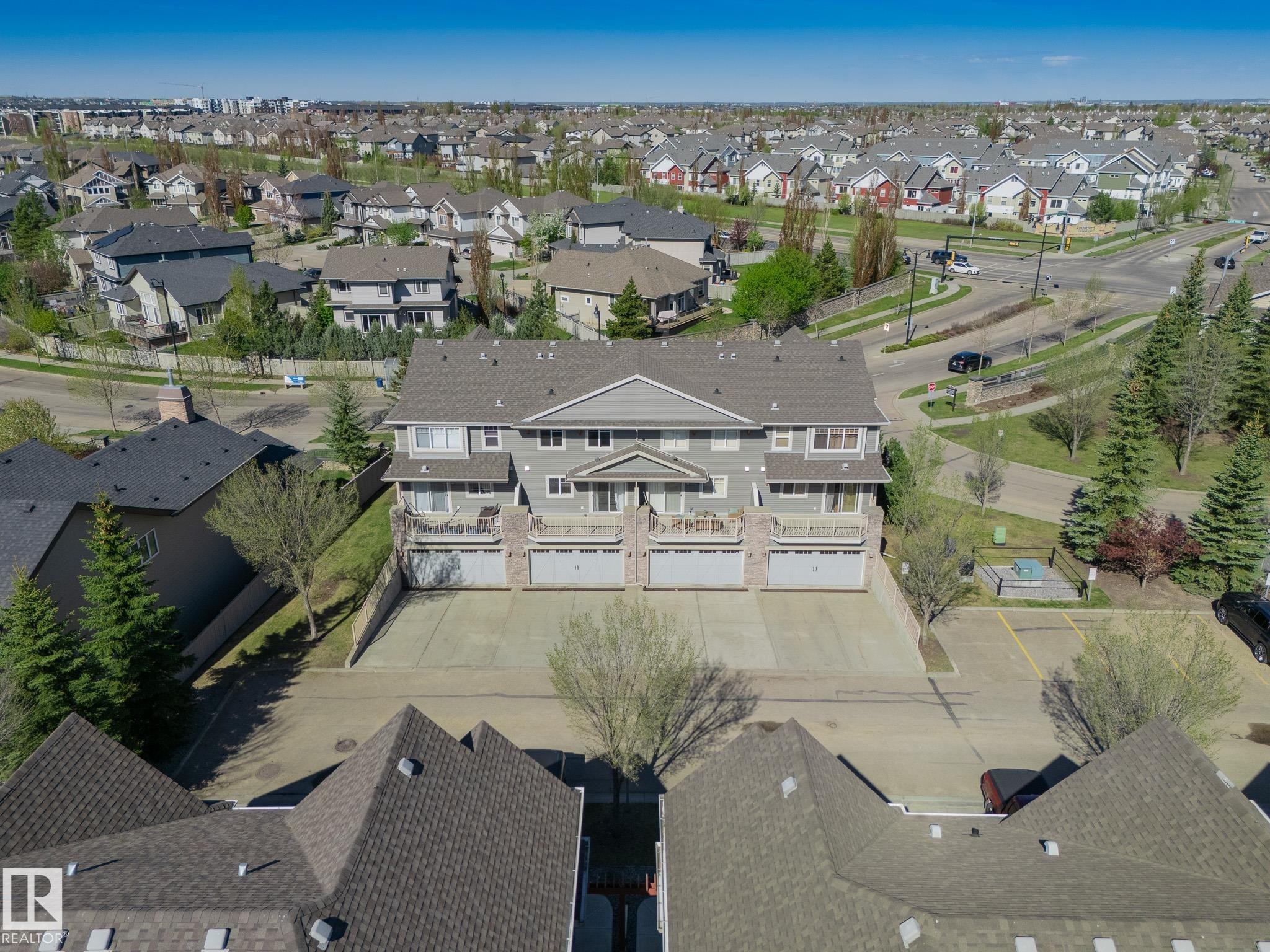 Multi-unit property featuring a neutral gray facade with white trim, multiple balconies with stone accents, individual ground-level garages, and a large paved parking area - 117 Callaghan Drive Sw, Edmonton, AB - Outdoor With View