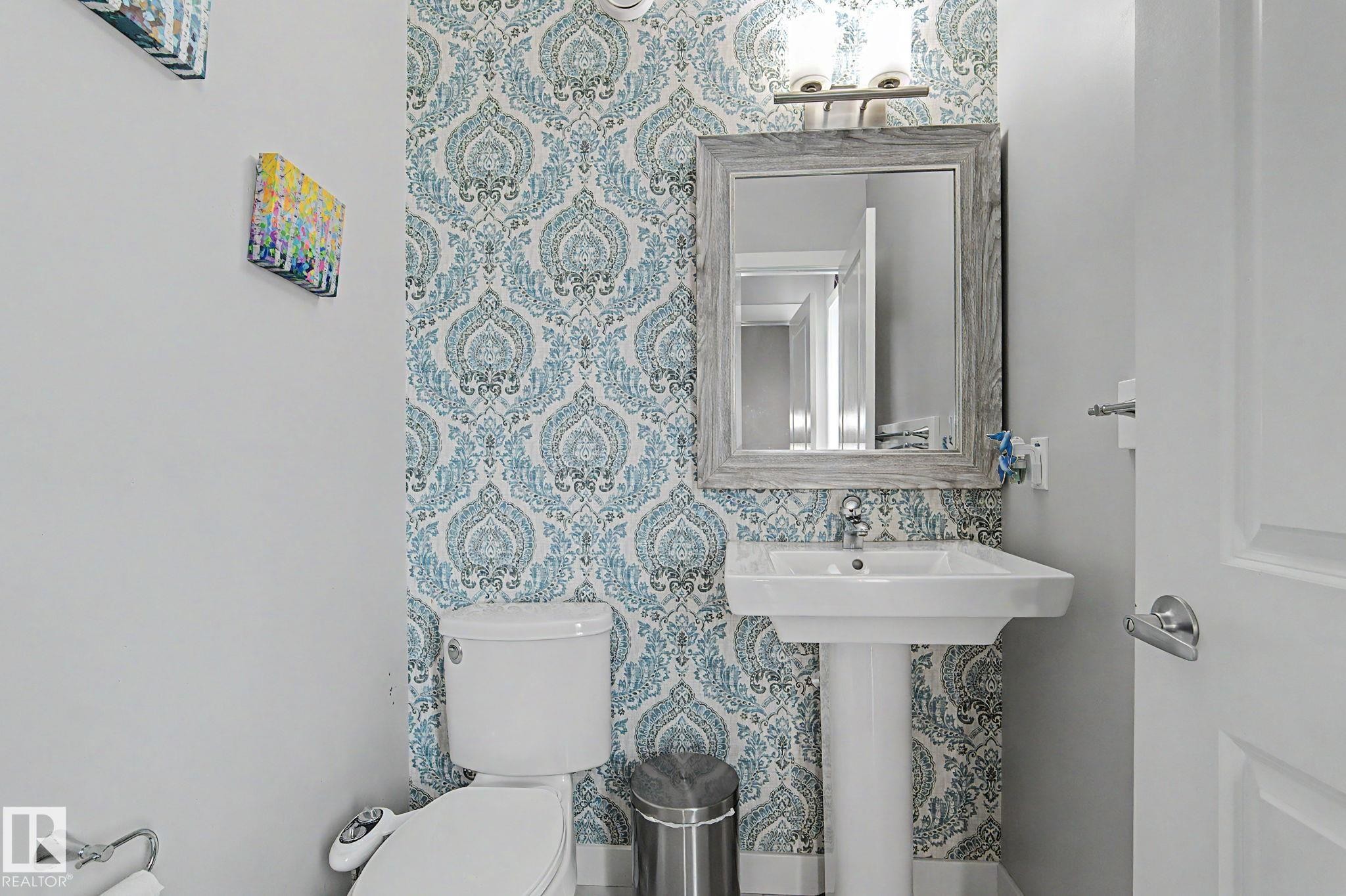 Pedestal sink with chrome faucet, framed mirror, and decorative wallpaper accent wall, complemented by a white toilet and overhead vanity lighting - 18144 76 Street Nw, Edmonton, AB - Indoor Photo Showing Bathroom
