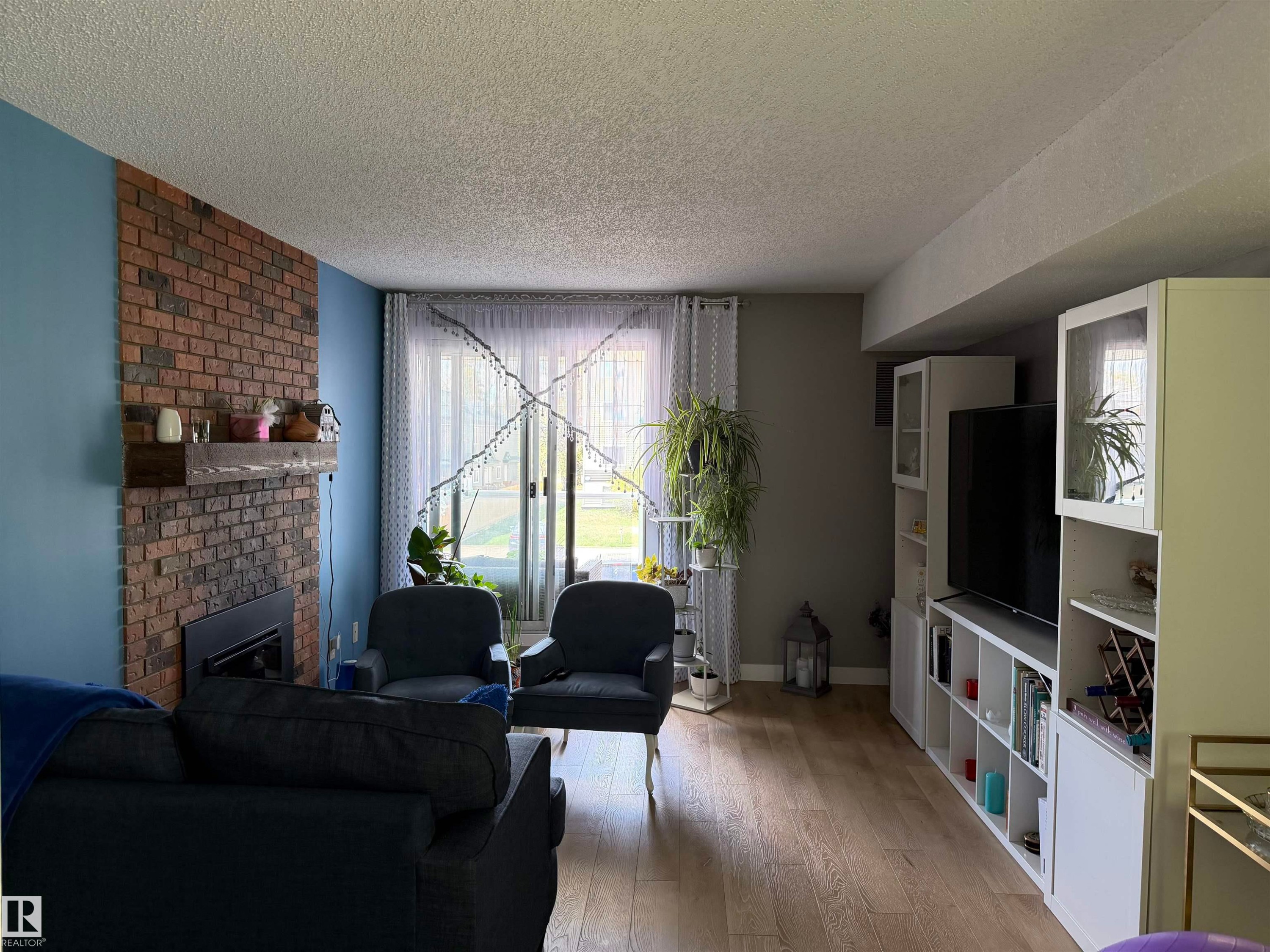 Living area featuring a brick-clad fireplace with a wood mantle, wood-finish flooring, and white built-in cabinetry - 205 10604 110 Avenue Nw, Edmonton, AB - Indoor Photo Showing Living Room With Fireplace
