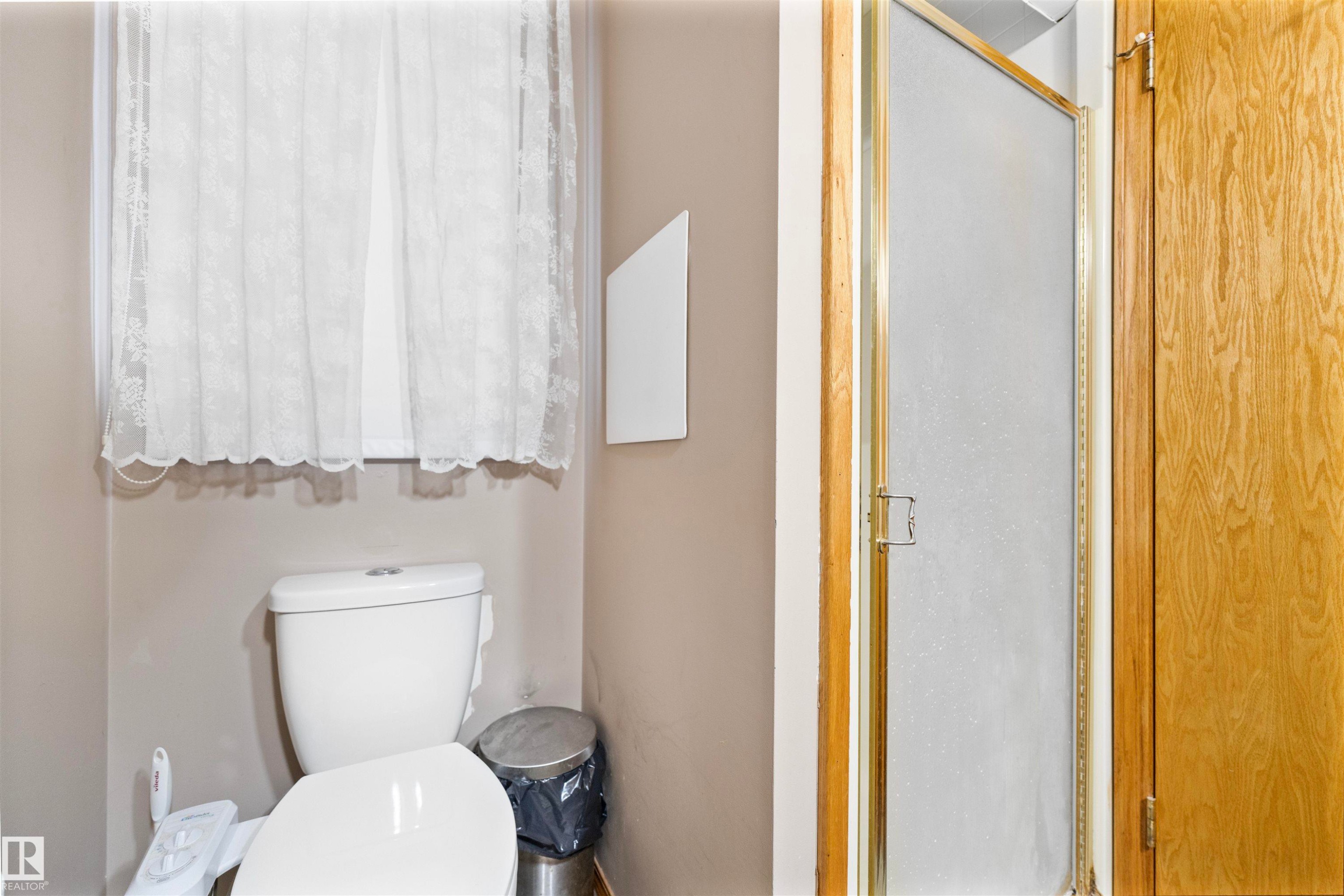 Compact bathroom featuring a white toilet, window with privacy curtain, and a shower with a frosted glass door and brass-tone framing - 4231 32 Avenue, Edmonton, AB - Indoor Photo Showing Bathroom