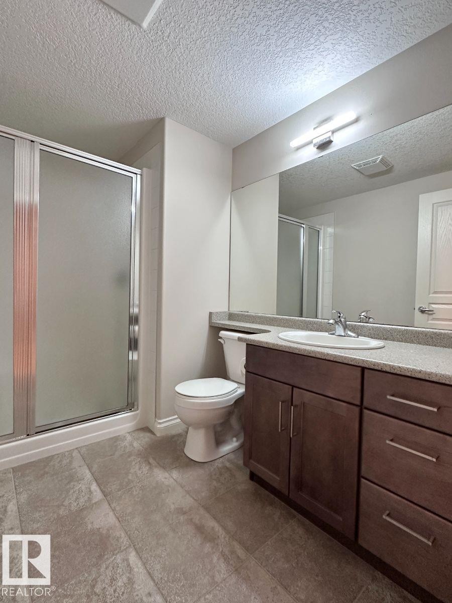 Bathroom featuring a glass-enclosed shower, a vanity with a white basin and chrome faucet, a large mirror, a toilet, and textured floor tiles - 820 Drysdale Run, Edmonton, AB - Indoor Photo Showing Bathroom