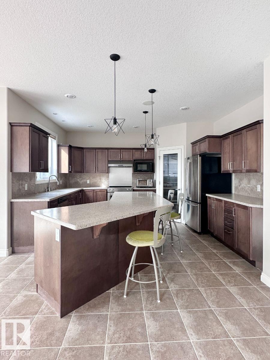 Spacious kitchen featuring an extended island with countertop seating, rich wood-finish cabinetry, tile flooring, modern pendant lighting, and recessed ceiling lights - 820 Drysdale Run, Edmonton, AB - Indoor Photo Showing Kitchen
