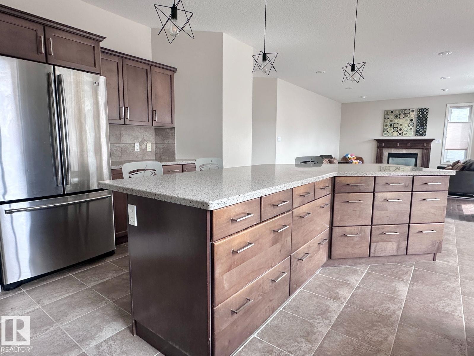 Spacious kitchen featuring a large central island with light-toned countertops, extensive wood cabinetry with modern hardware, stainless steel refrigerator, and tiled flooring - 820 Drysdale Run, Edmonton, AB - Indoor Photo Showing Kitchen With Upgraded Kitchen
