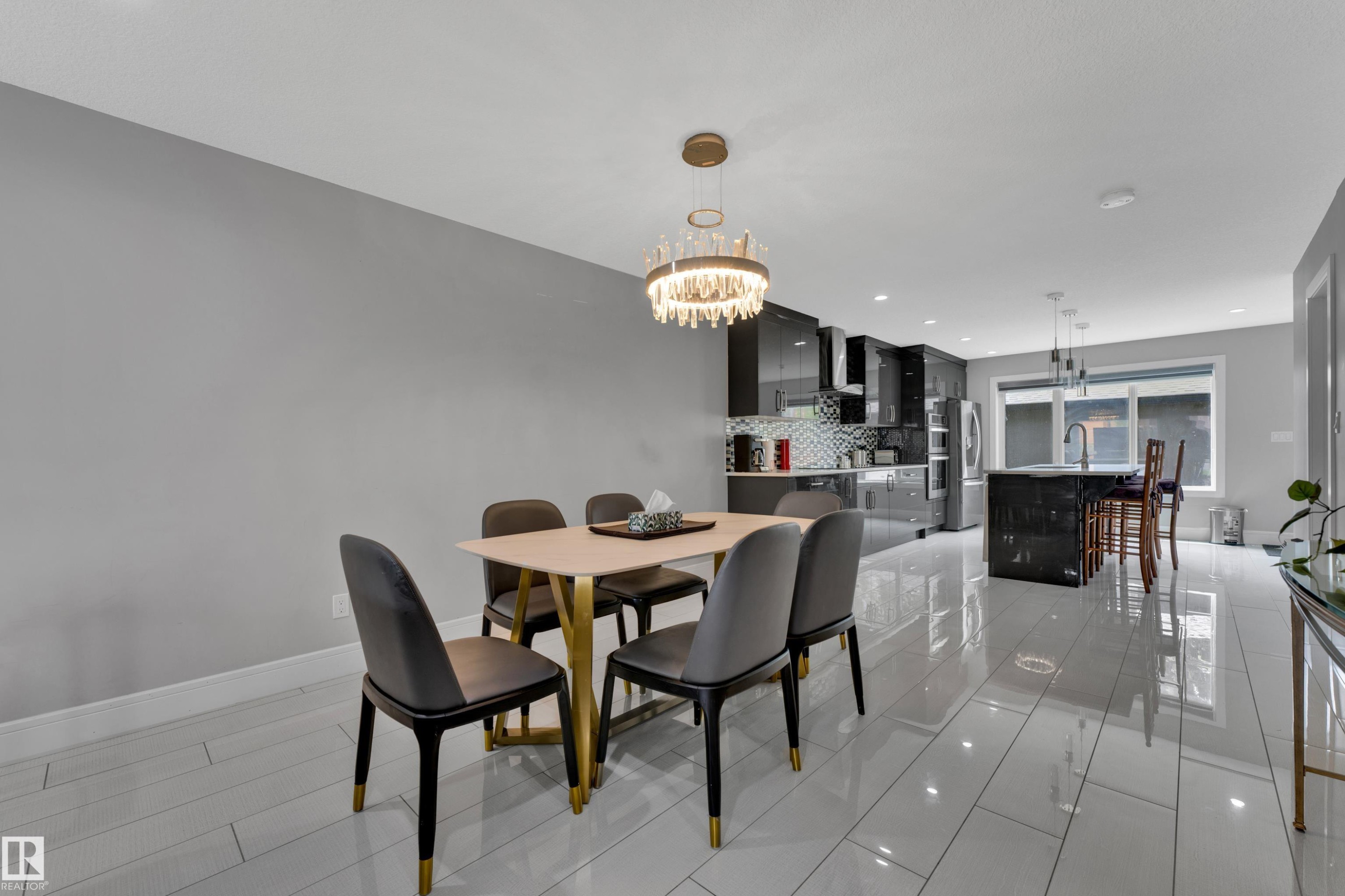 Open-concept dining area with a contemporary chandelier, tile flooring, and a sightline to the kitchen featuring glossy dark cabinetry, an island with seating, and stainless steel appliances - 9329 Connors Road, Edmonton, AB - Indoor Photo Showing Dining Room