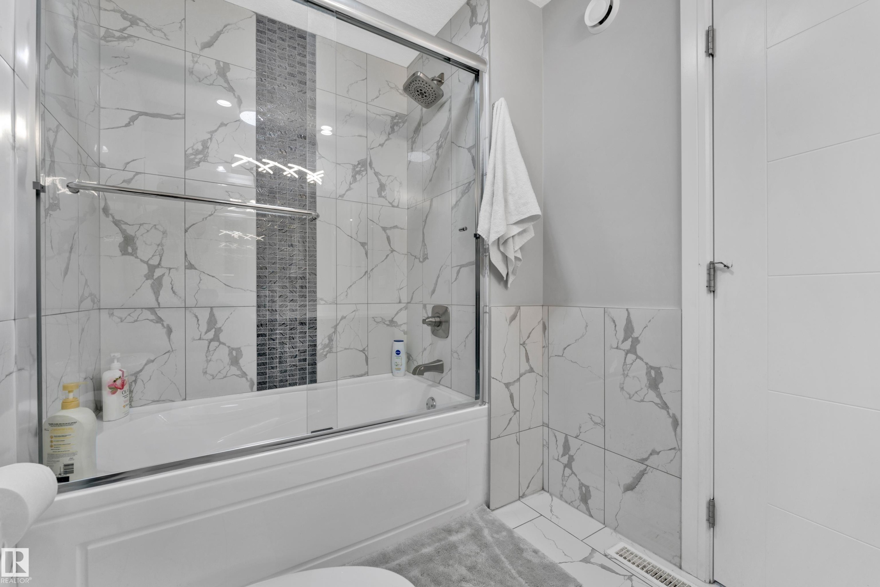 Bathtub with glass sliding doors, featuring a tiled surround with a vertical mosaic accent - 9329 Connors Road, Edmonton, AB - Indoor Photo Showing Bathroom