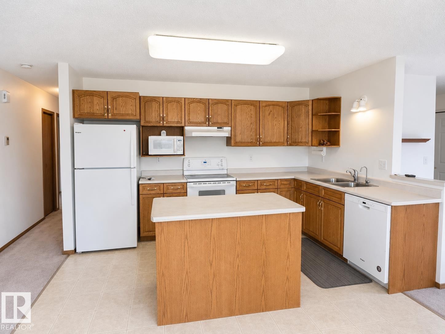 Kitchen featuring wood cabinetry, white appliances, a center island with a light-toned countertop, a double basin sink, and tile flooring - 4005 55 Avenue, Whitecourt, AB - Indoor Photo Showing Kitchen With Double Sink