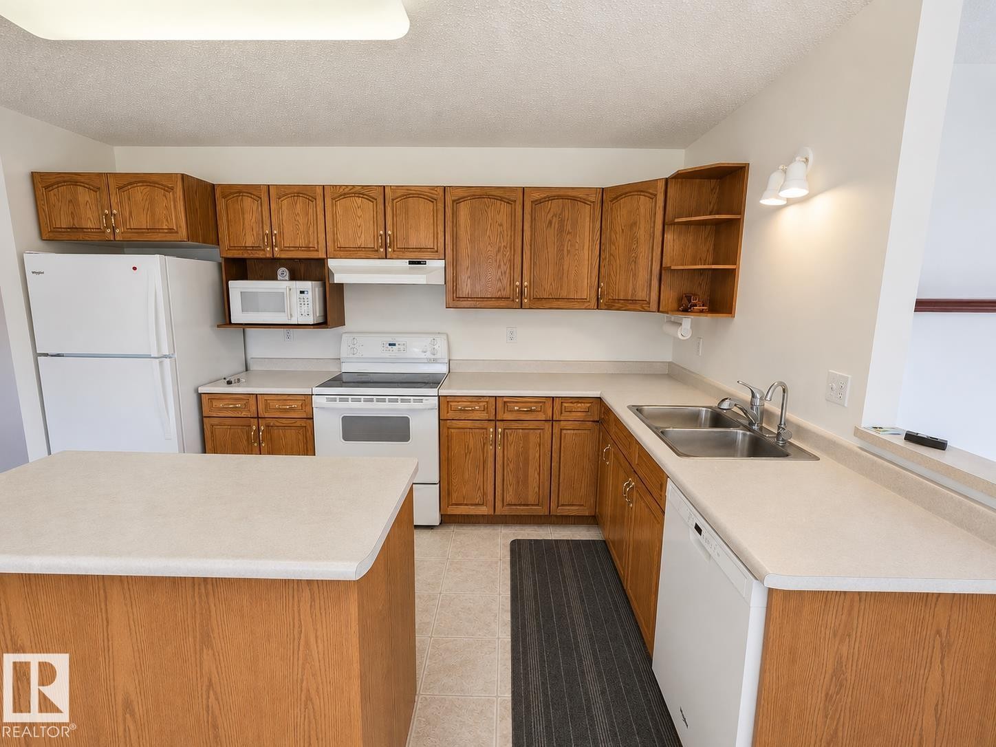 Bright kitchen featuring wood-finish cabinetry, white appliances, a double basin stainless steel sink, and light-toned countertops - 4005 55 Avenue, Whitecourt, AB - Indoor Photo Showing Kitchen With Double Sink