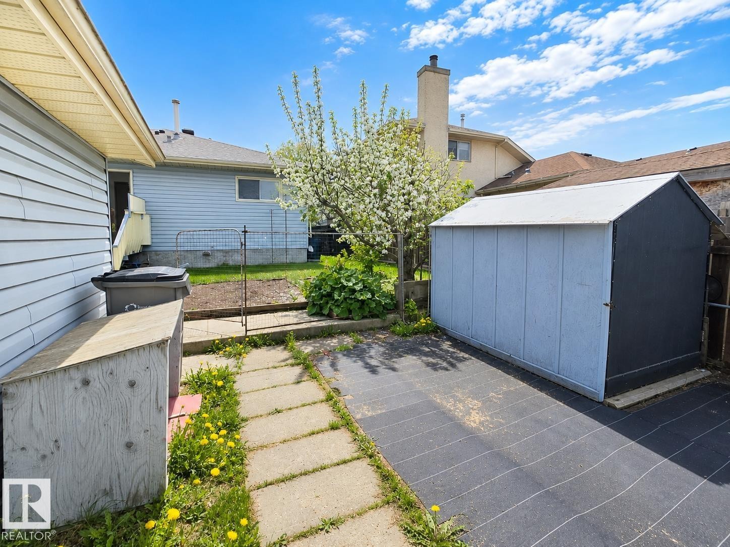 Backyard space featuring a storage shed with light blue and dark gray siding, a blossoming tree, and a stone paver pathway - 4005 55 Avenue, Whitecourt, AB - Outdoor With Exterior