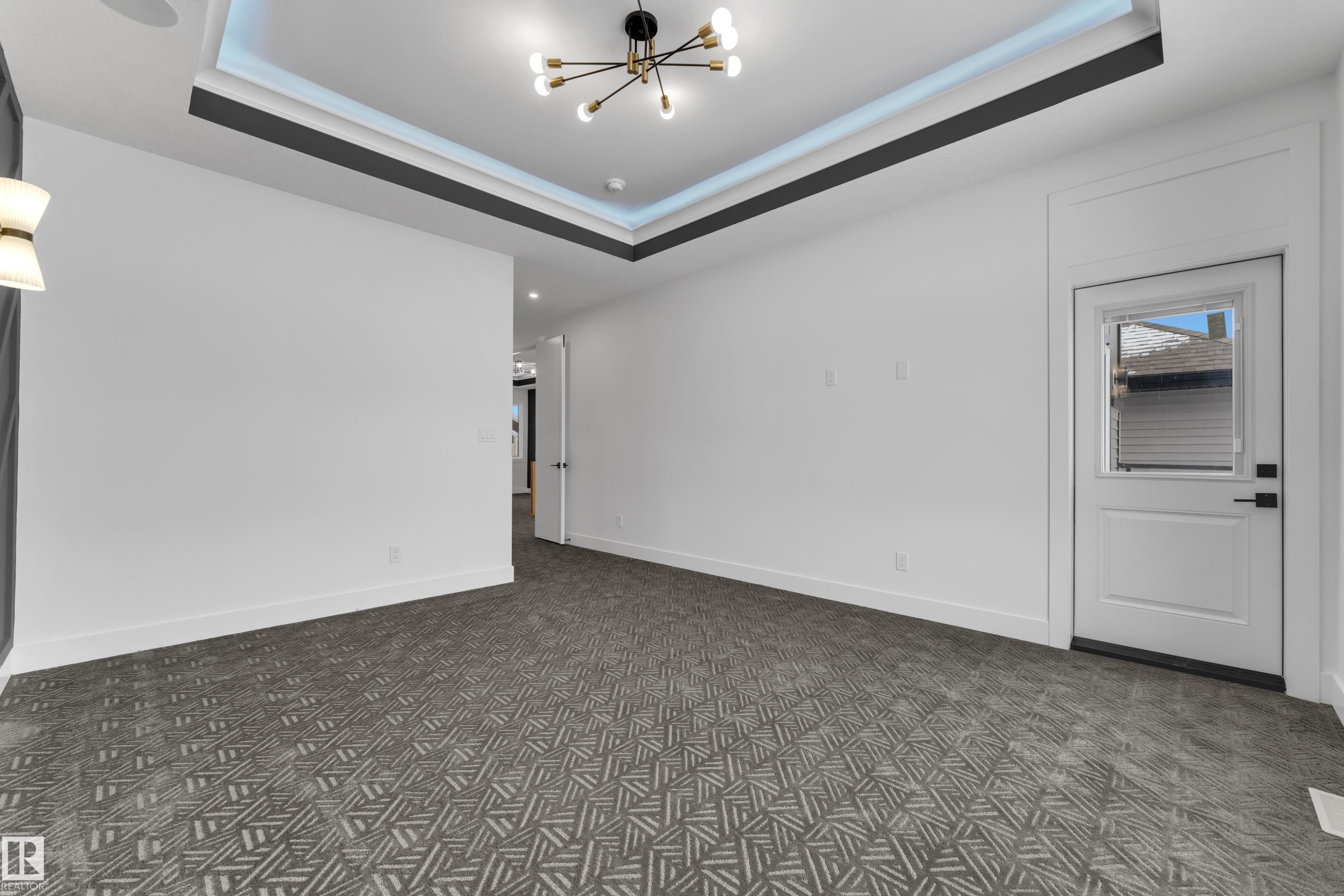 Expansive room featuring patterned carpeting, a tray ceiling with integrated lighting, a contemporary chandelier, white walls, and a white exterior door with a window - 2027 156 Avenue, Edmonton, AB - Indoor Photo Showing Other Room