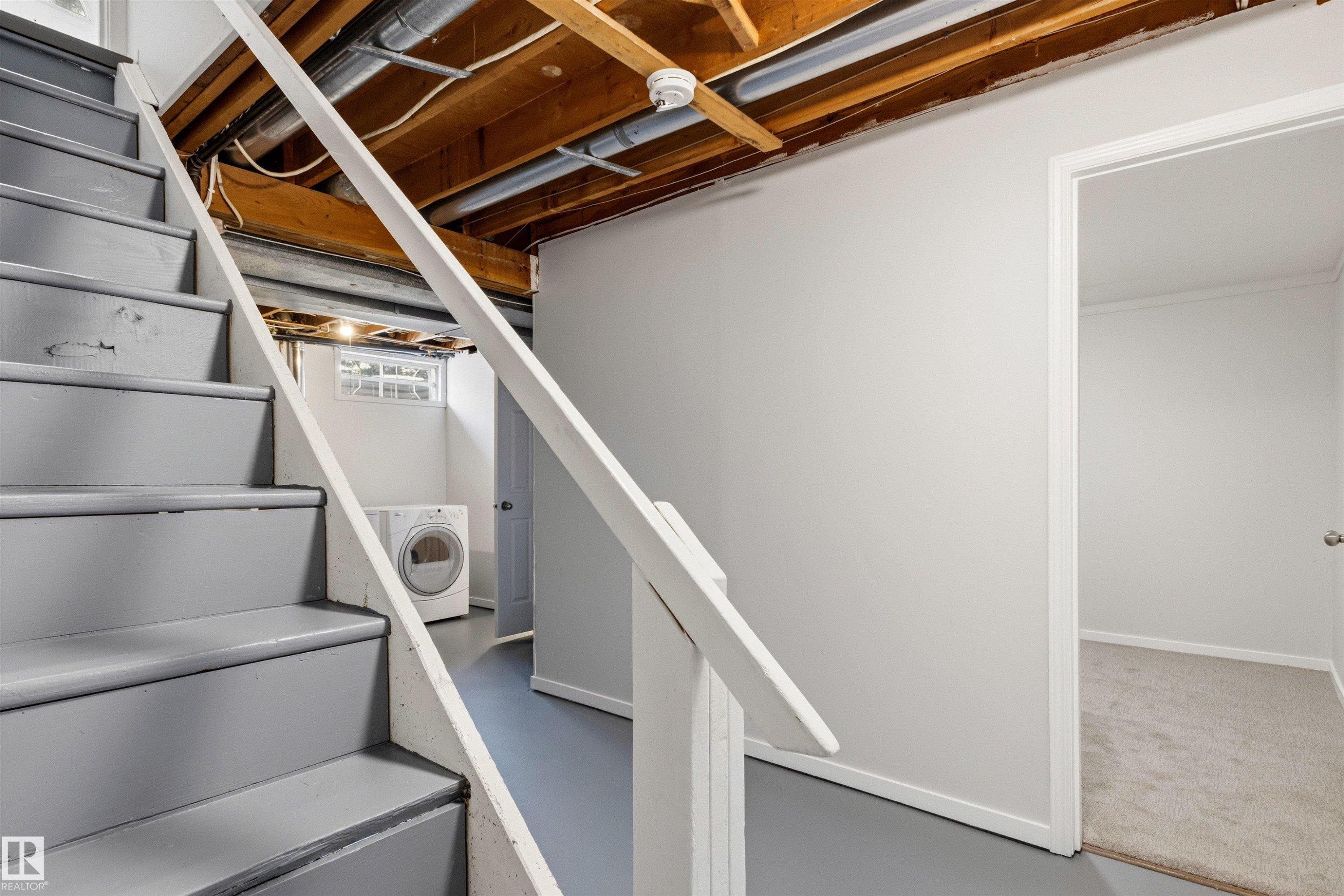 Painted stairs with a white handrail lead to a finished lower level featuring light gray walls, a window with glass block, and a carpeted room beyond an open doorway - 707 Clareview Road, Edmonton, AB - Indoor Photo Showing Laundry Room
