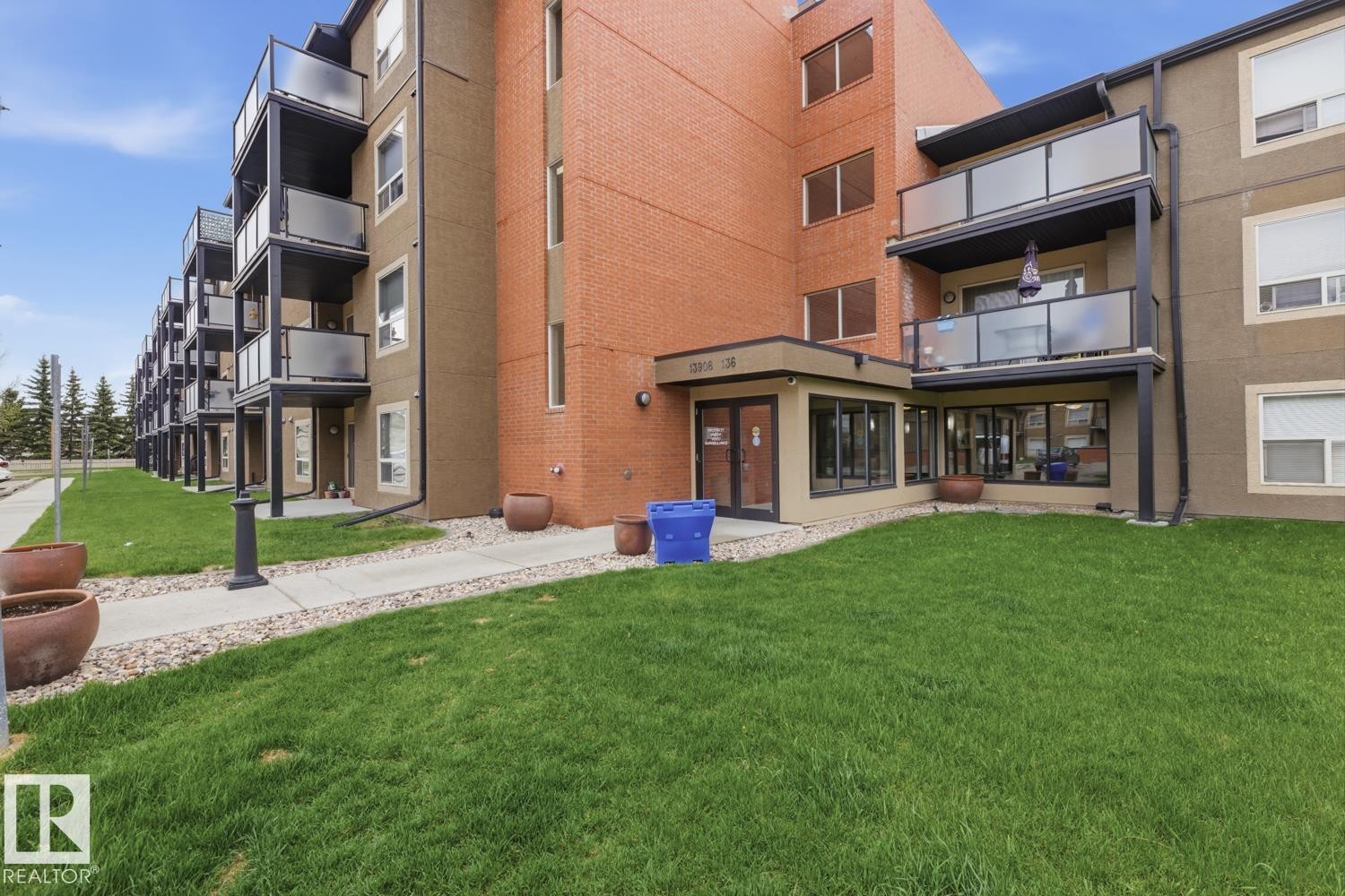 Modern multi-story building featuring a brick and stucco exterior, individual balconies with glass railings, a ground-level entrance with extensive fenestration, a concrete walkway, and surrounding green space - #312 13908 136 St Nw, Edmonton, AB - Outdoor With Balcony
