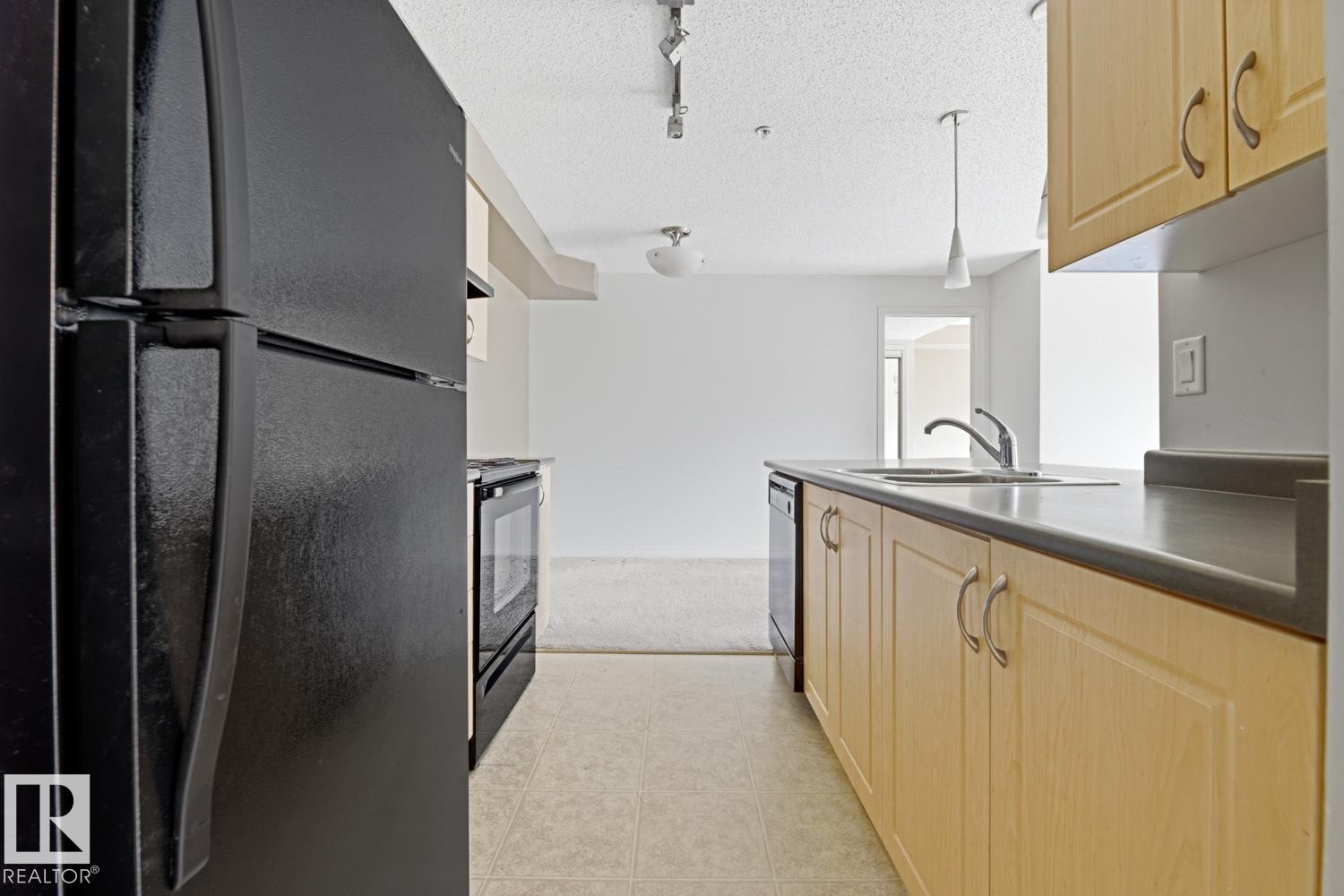 Galley kitchen featuring light wood-finish cabinetry, black countertops, a stainless steel sink with a chrome faucet, and black appliances including a refrigerator, range, and dishwasher - #312 13908 136 St Nw, Edmonton, AB - Indoor Photo Showing Kitchen