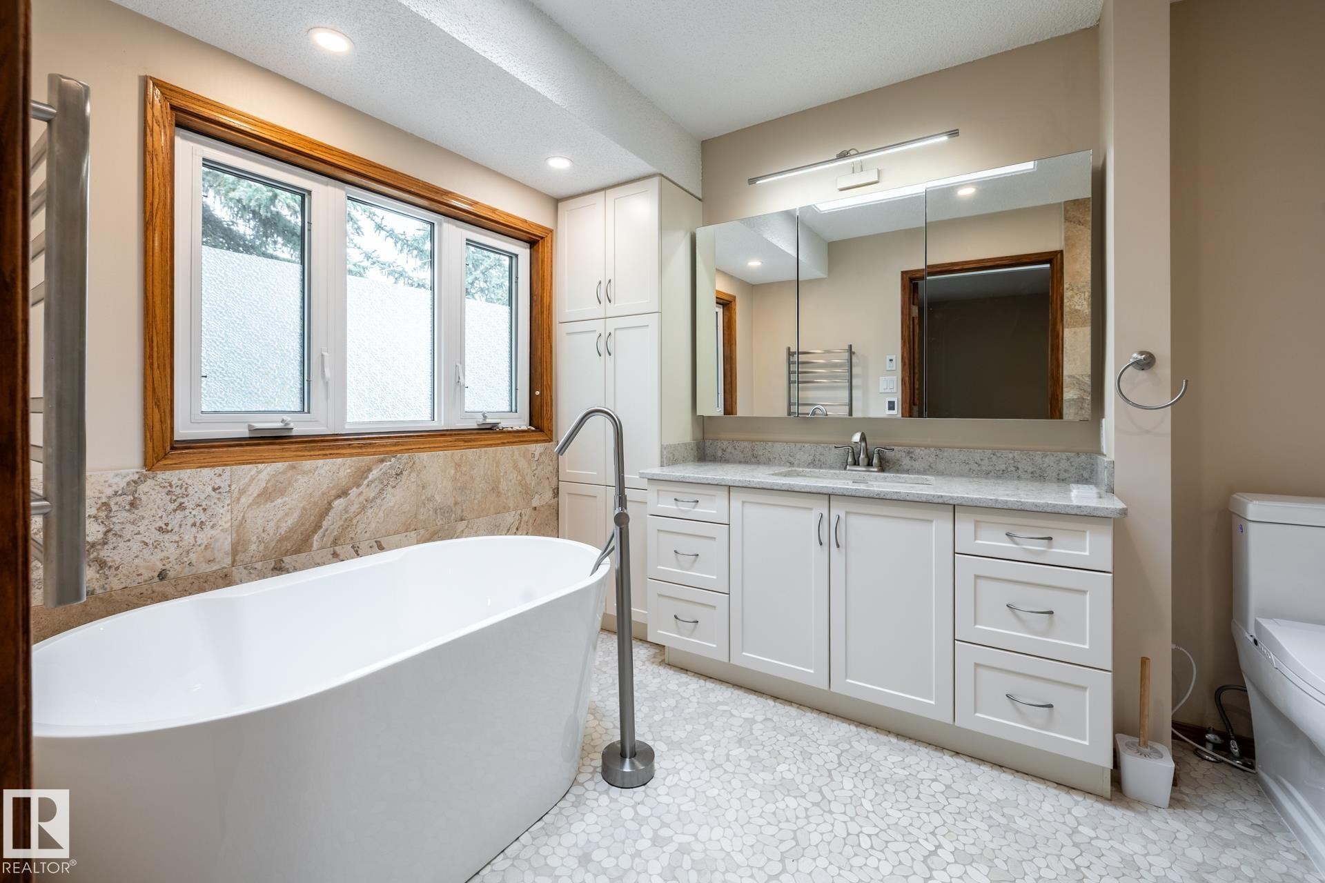 Freestanding soaking tub with a floor-mounted faucet, complemented by a large three-pane window with wood-finish trim and natural stone-look tile surround - 4632 151 Street, Edmonton, AB - Indoor Photo Showing Bathroom