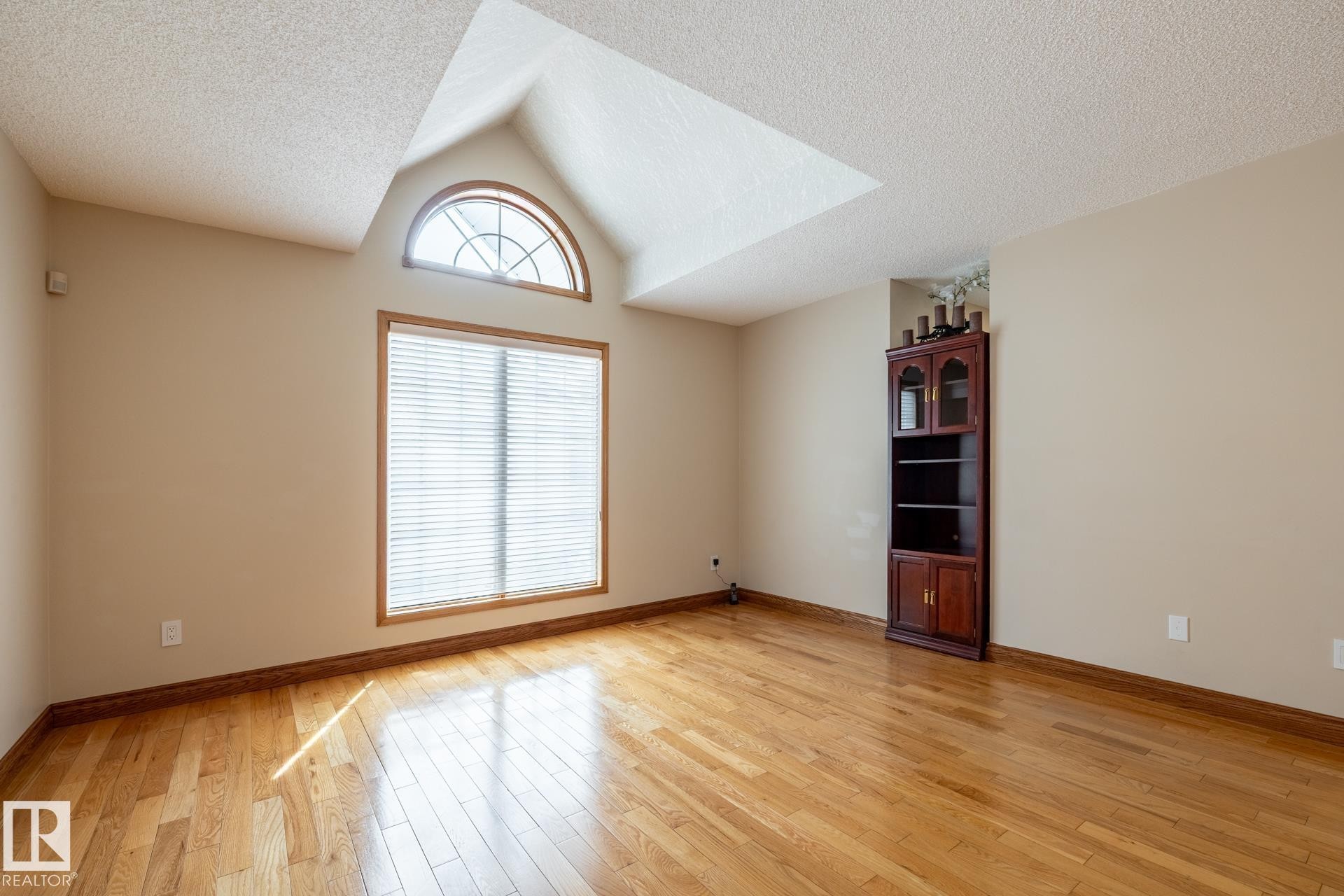 Hardwood flooring throughout featuring a large picture window with blinds, an arched transom window, and a built-in corner cabinet - 4632 151 Street, Edmonton, AB - Indoor Photo Showing Other Room