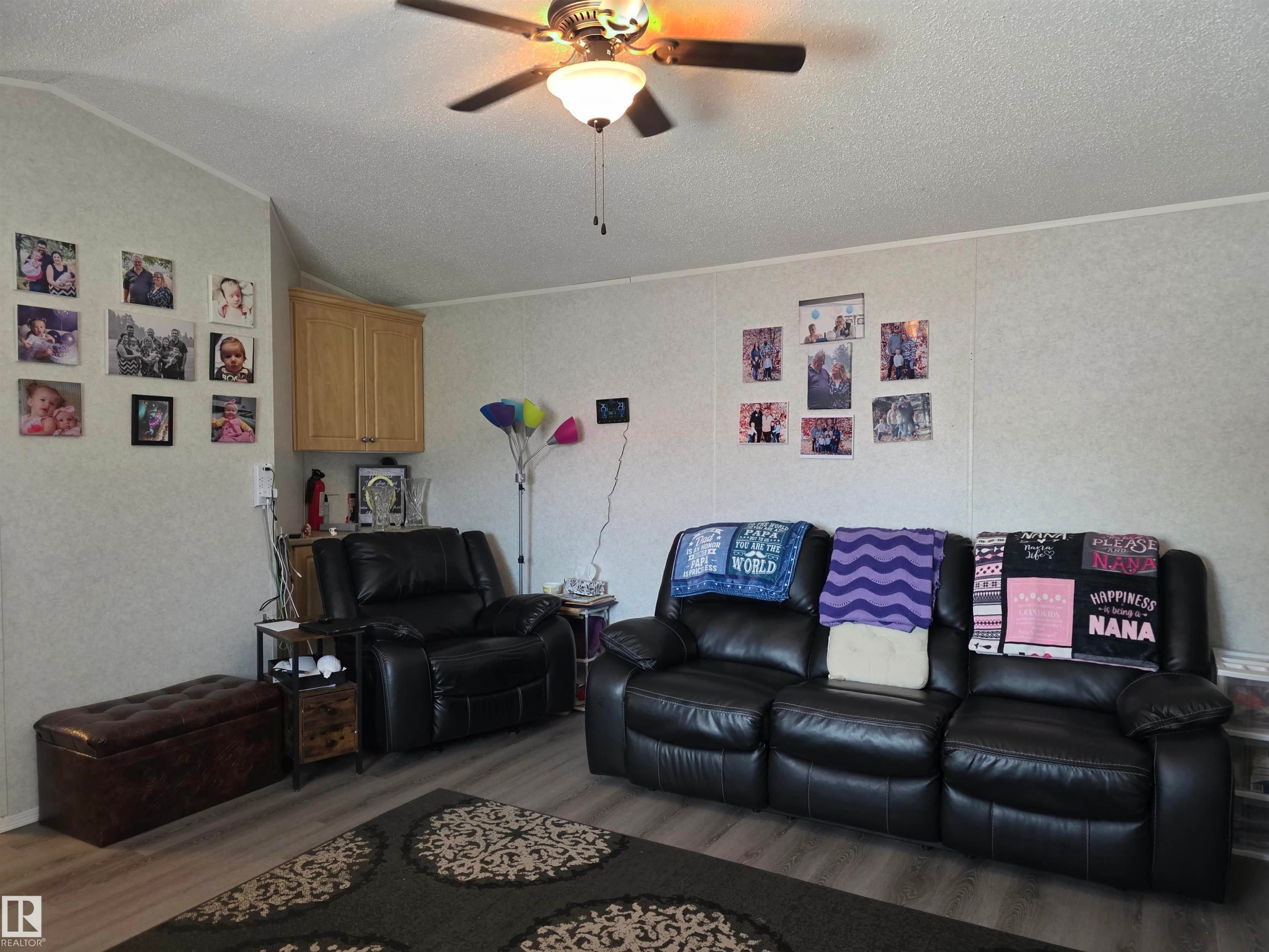 Living area featuring wood-finish flooring, a ceiling fan with integrated lighting, textured wall panels, and a corner-mounted wood-finish cabinet - 5012 53 Ave, Breton, AB
