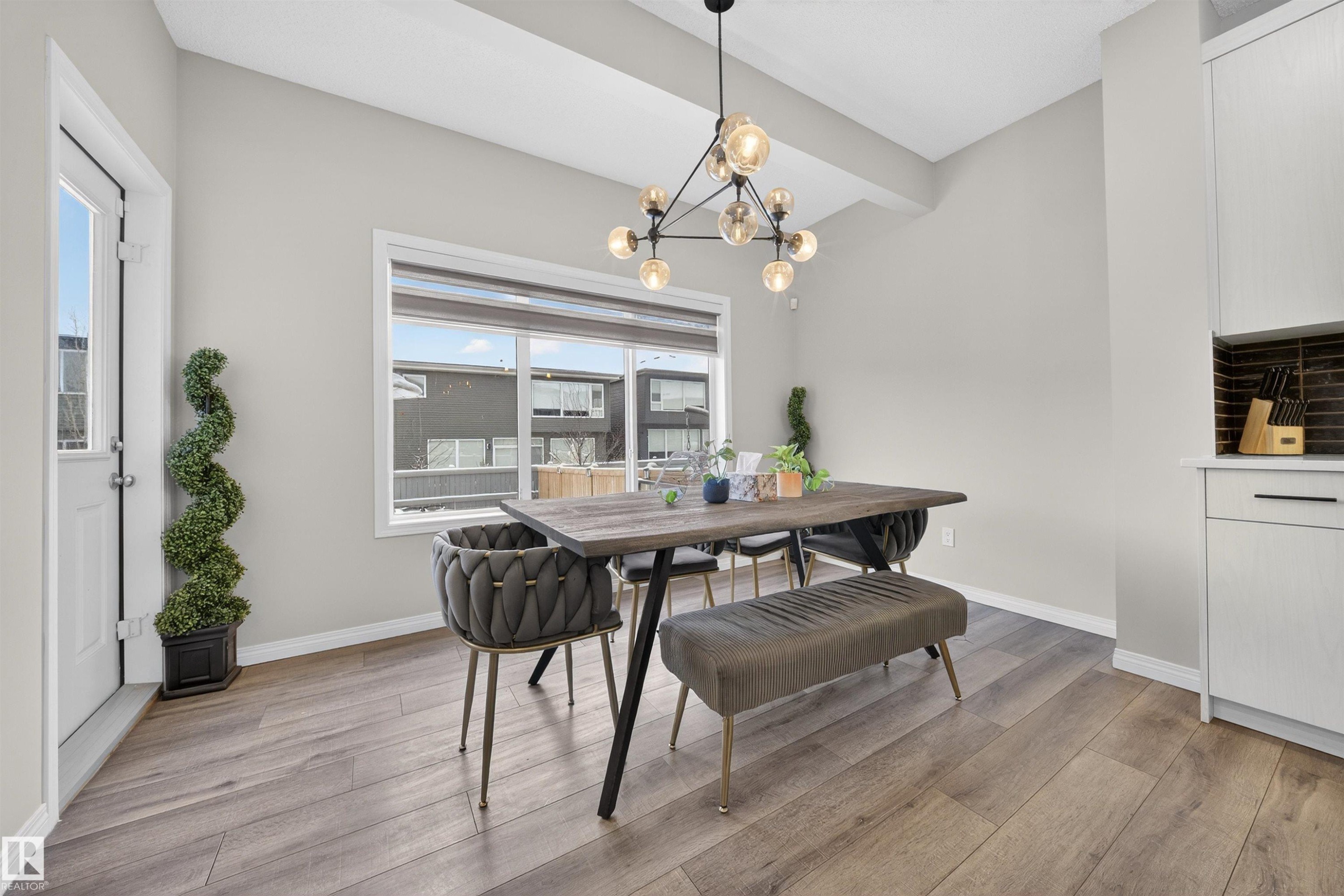 Spacious dining area featuring wood-finish flooring, a contemporary chandelier, large window with natural light, a white exterior door, and built-in cabinetry with a dark tile backsplash - 6085 King Landing, Edmonton, AB - Indoor Photo Showing Dining Room