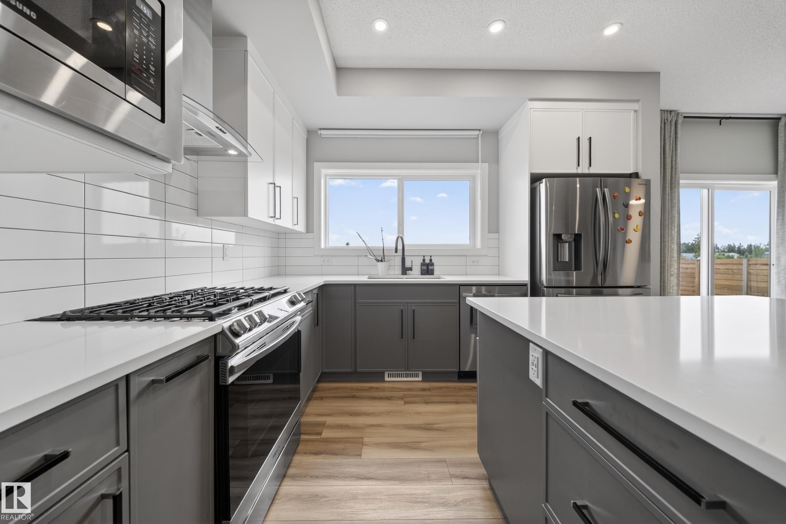 Kitchen featuring an expansive island with a white countertop, wood-finish flooring, white subway tile backsplash, stainless steel appliances, and a window above the sink - 3337 Kulay Way, Edmonton, AB - Indoor Photo Showing Kitchen With Upgraded Kitchen