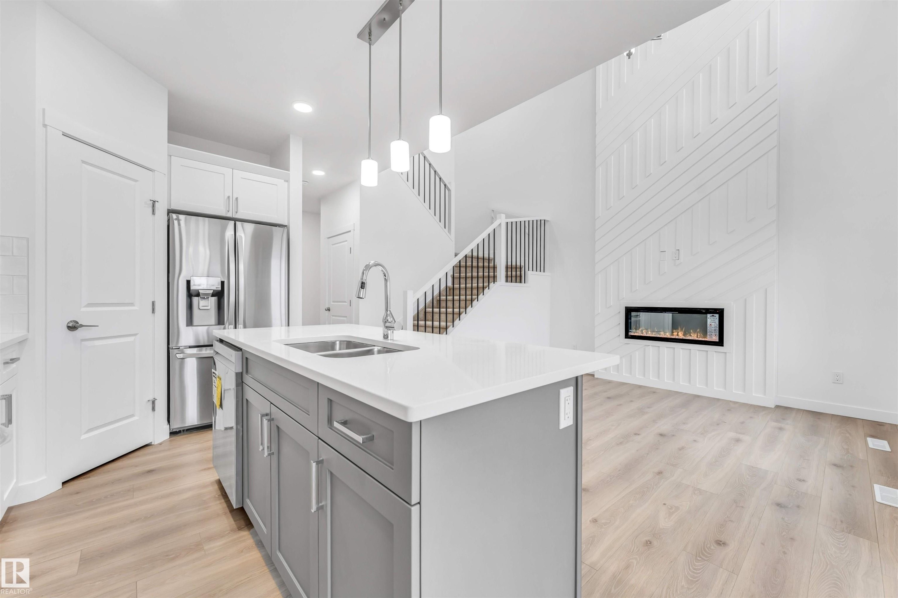 Open-concept kitchen and living area featuring a large island with sink, stainless steel appliances, wood-finish flooring, and a modern fireplace with a textured accent wall - 1972 210 Street, Edmonton, AB - Indoor Photo Showing Kitchen With Double Sink With Upgraded Kitchen