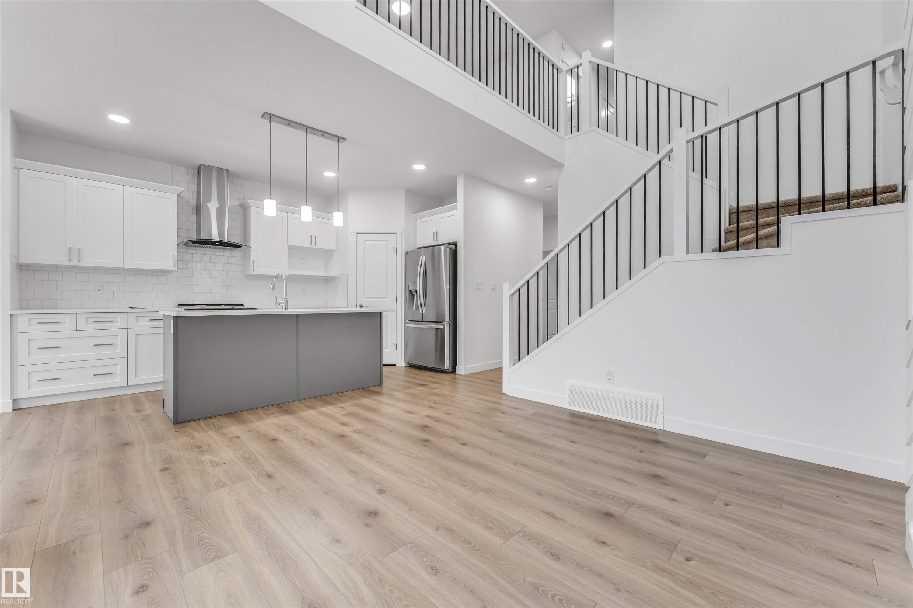 Open-concept living area featuring light wood-finish flooring, a contemporary kitchen with white cabinetry, a gray kitchen island, stainless steel appliances, and a two-story staircase with black metal railings - 1972 210 Street, Edmonton, AB - Indoor Photo Showing Kitchen With Upgraded Kitchen