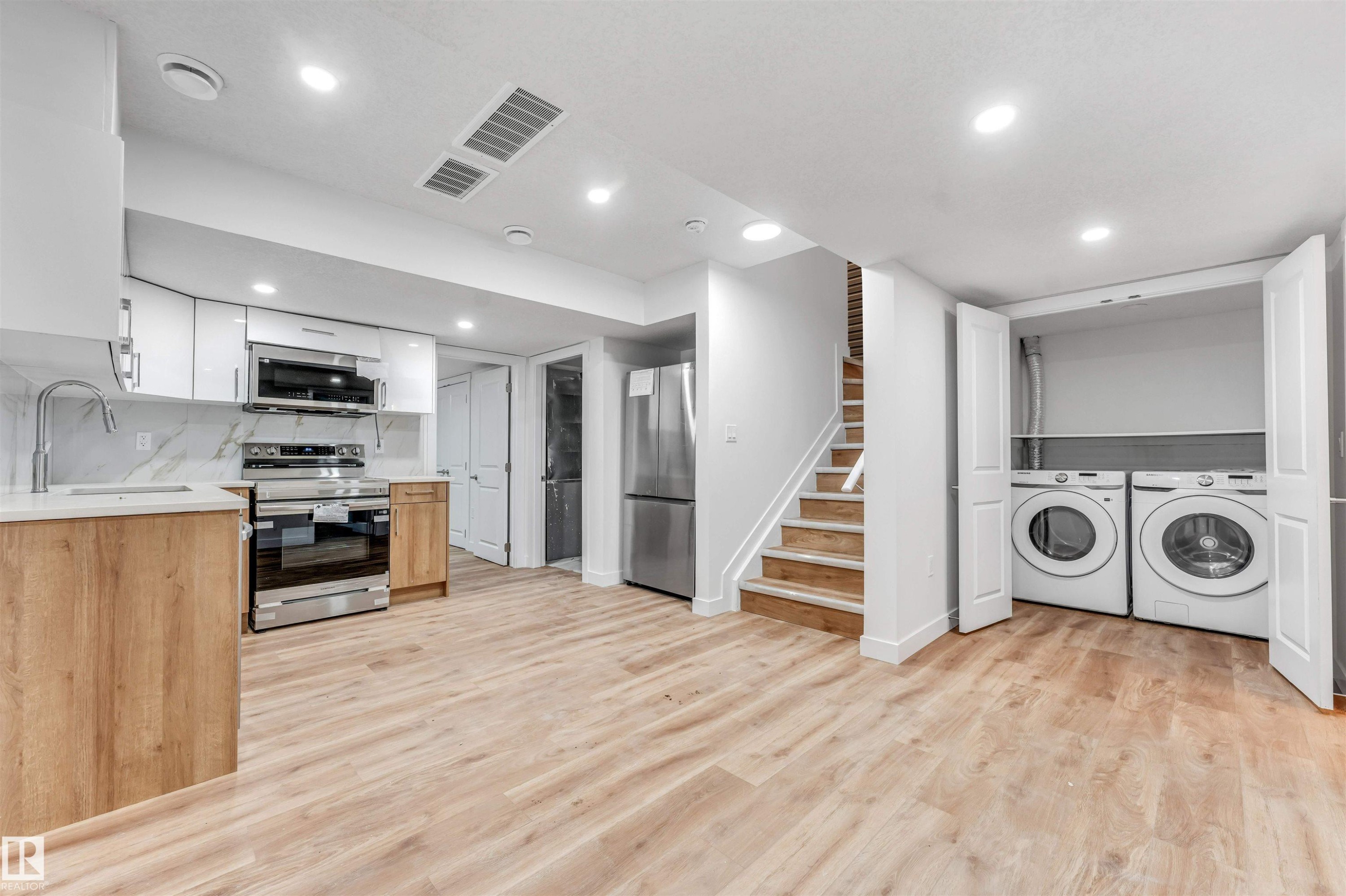 Open-concept living area featuring a modern kitchen with stainless steel appliances, light wood-finish flooring, recessed lighting, and a dedicated laundry closet with a stackable washer and dryer - 1972 210 Street, Edmonton, AB - Indoor Photo Showing Laundry Room