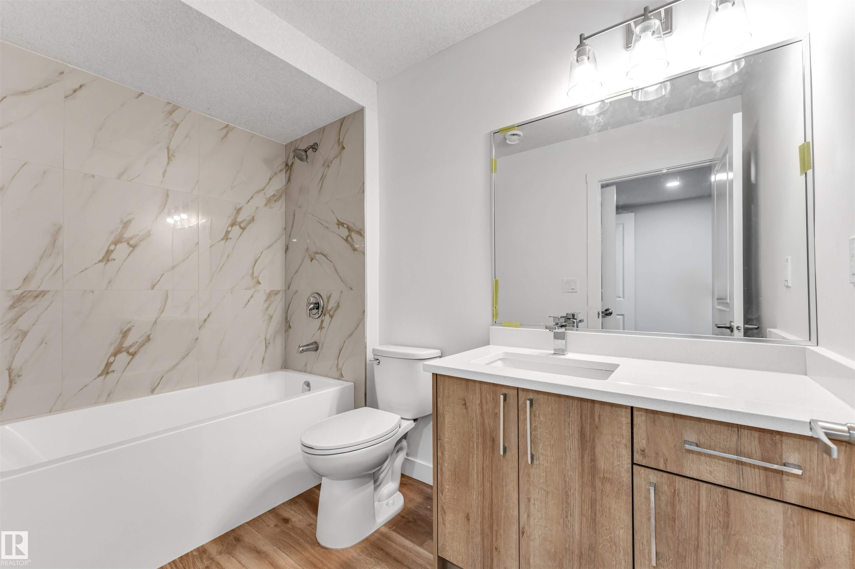 Bathroom featuring a white bathtub with full-height marble-look tile surround, a wood-finish vanity with a white countertop and undermount sink, and a large mirror with a four-light fixture - 1972 210 Street, Edmonton, AB - Indoor Photo Showing Bathroom
