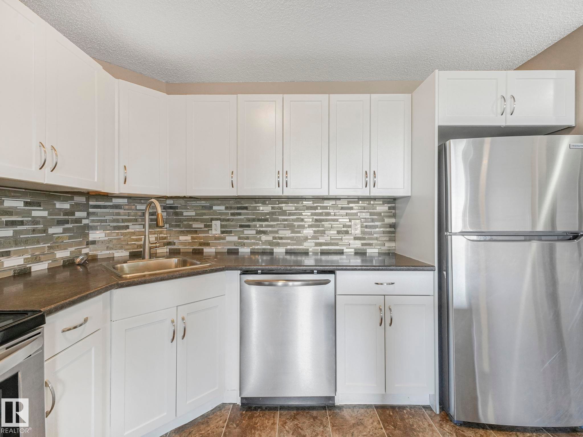 Kitchen featuring white cabinetry, dark countertops, a mosaic tile backsplash, stainless steel appliances, and wood-finish flooring - 322 1520 Hammond Gate Nw, Edmonton, AB - Indoor Photo Showing Kitchen With Upgraded Kitchen