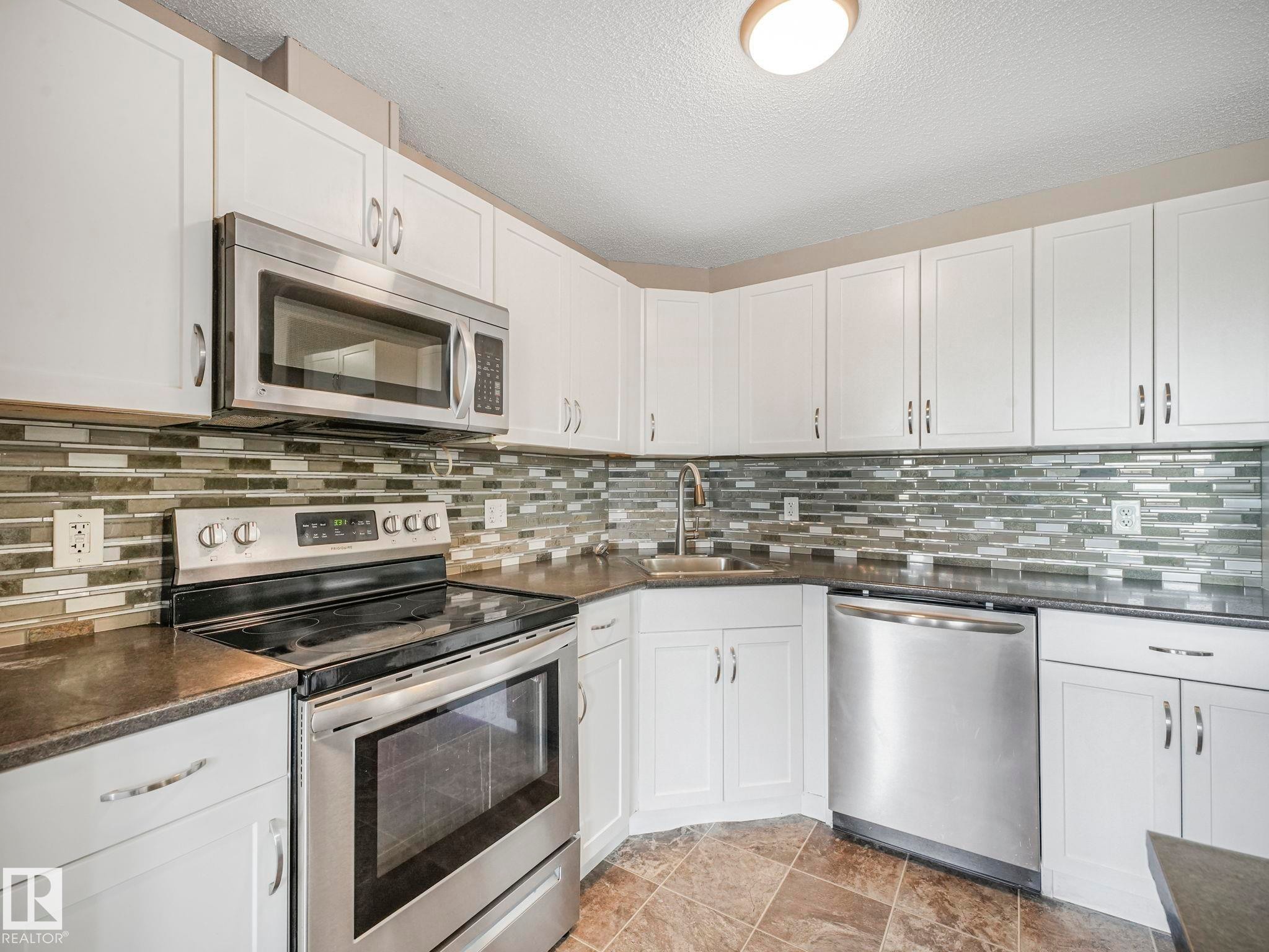 Modern kitchen featuring white cabinetry, stainless steel appliances, dark countertops, a mosaic tile backsplash, and tile flooring - 322 1520 Hammond Gate Nw, Edmonton, AB - Indoor Photo Showing Kitchen With Upgraded Kitchen