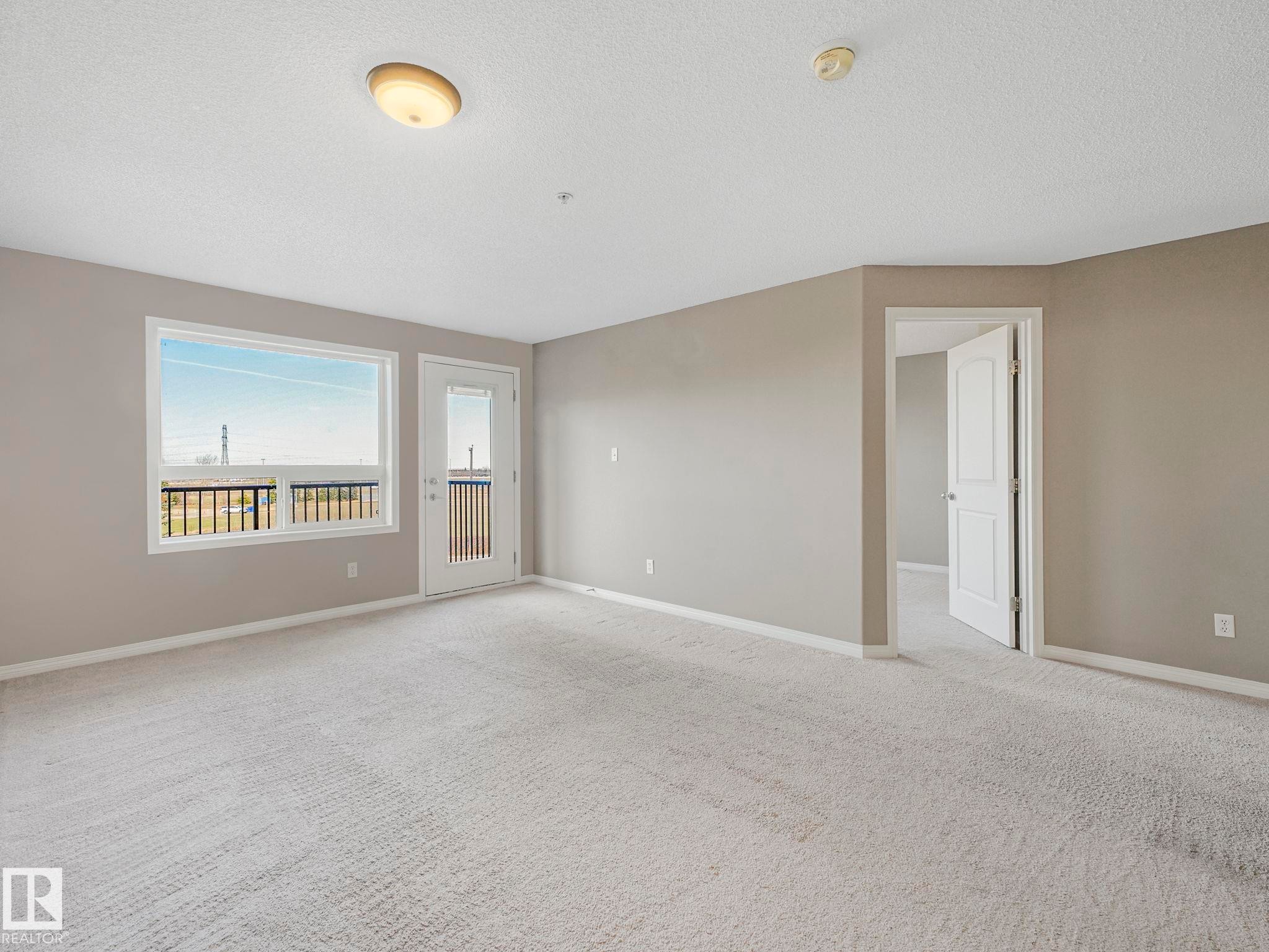 Carpeted living space featuring a window with a black railing, a door with glass panels, painted walls, and white trim - 322 1520 Hammond Gate Nw, Edmonton, AB - Indoor Photo Showing Other Room