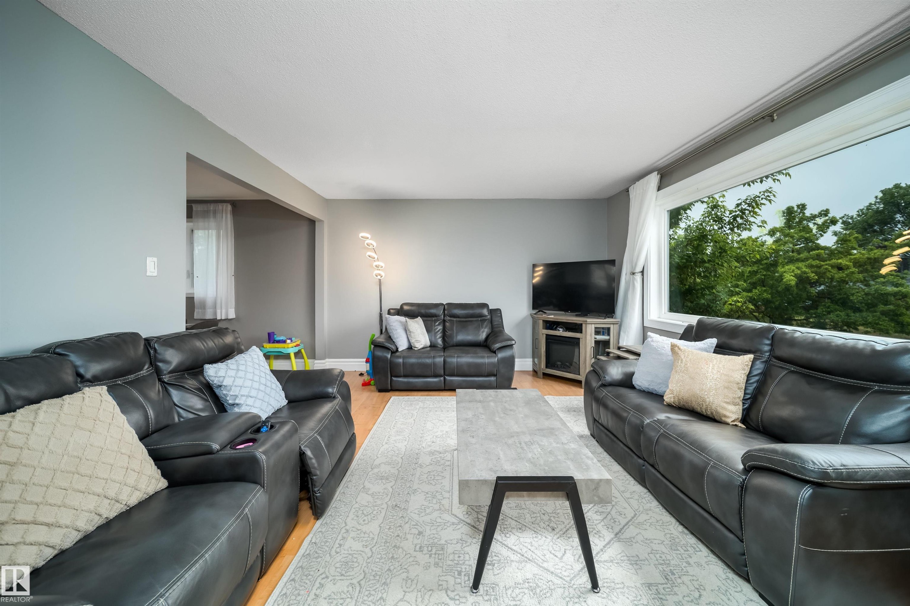 Spacious living area featuring light gray wall paint, wood-finish flooring, and a large picture window - 8512 142A Avenue, Edmonton, AB