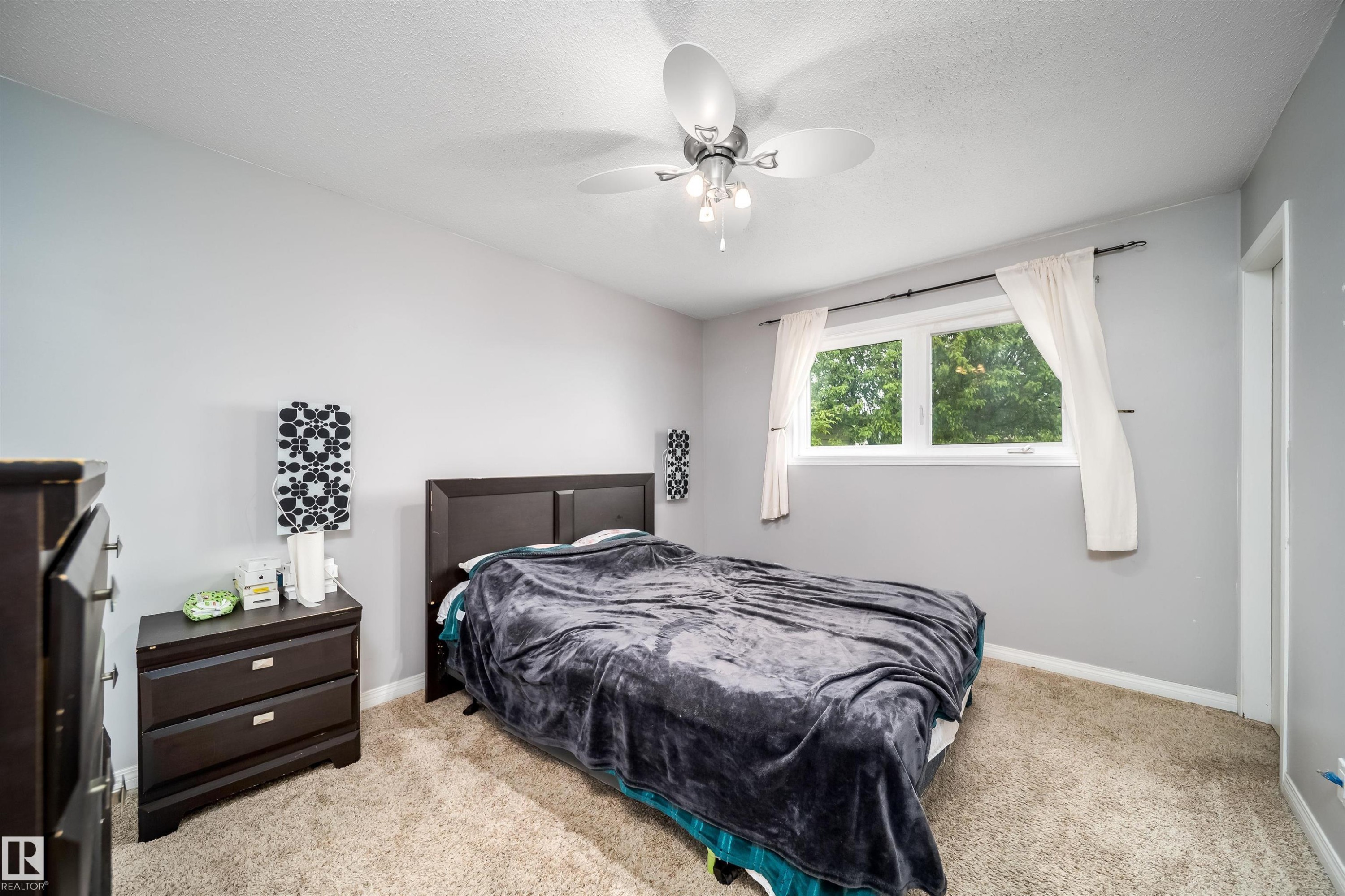 Carpeted bedroom featuring a large window, a ceiling fan with integrated lighting, and a solid-panel door - 8512 142A Avenue, Edmonton, AB