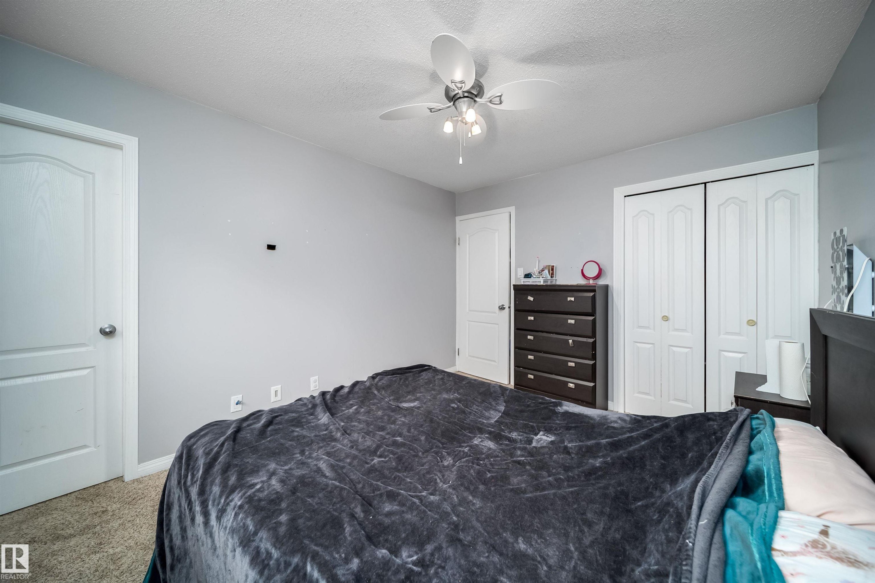 Carpeted bedroom featuring light gray walls, white trim, a built-in ceiling fan with light fixture, and white bi-fold closet doors - 8512 142A Avenue, Edmonton, AB