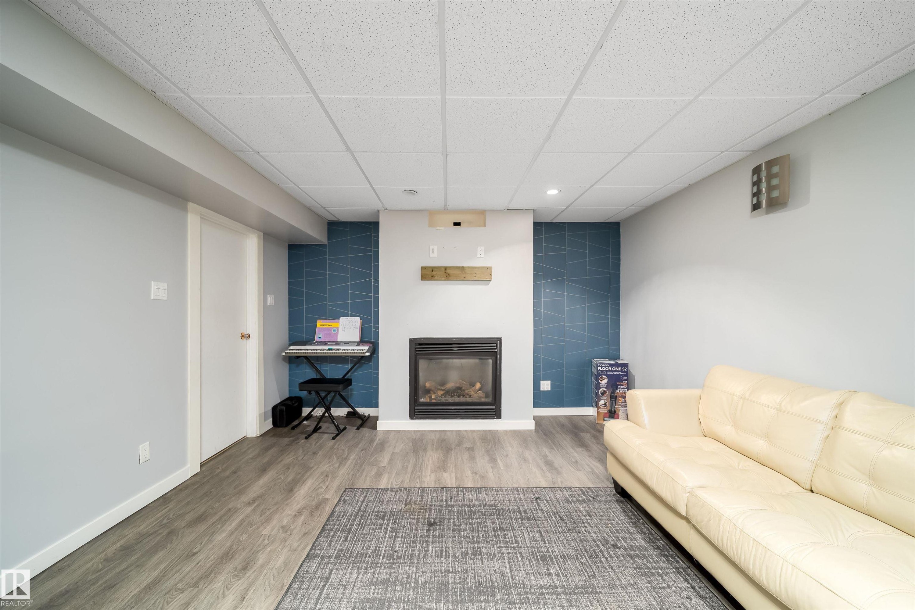 Living area featuring a built-in fireplace with a wood mantel, wood-finish flooring, a drop ceiling, and a distinct accent wall - 8512 142A Avenue, Edmonton, AB