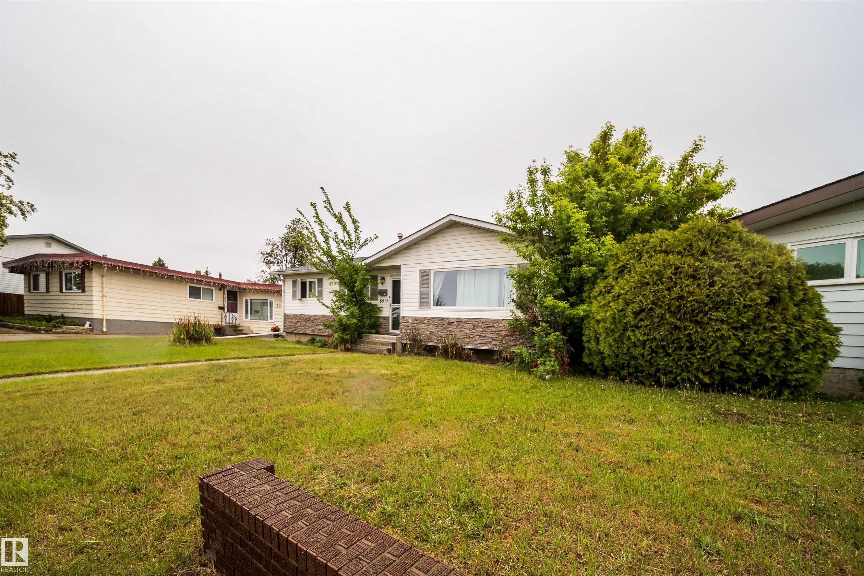 Expansive front lawn with mature landscaping, a brick garden wall, and a single-story residence featuring white siding and a stone-clad foundation - 8512 142A Avenue, Edmonton, AB