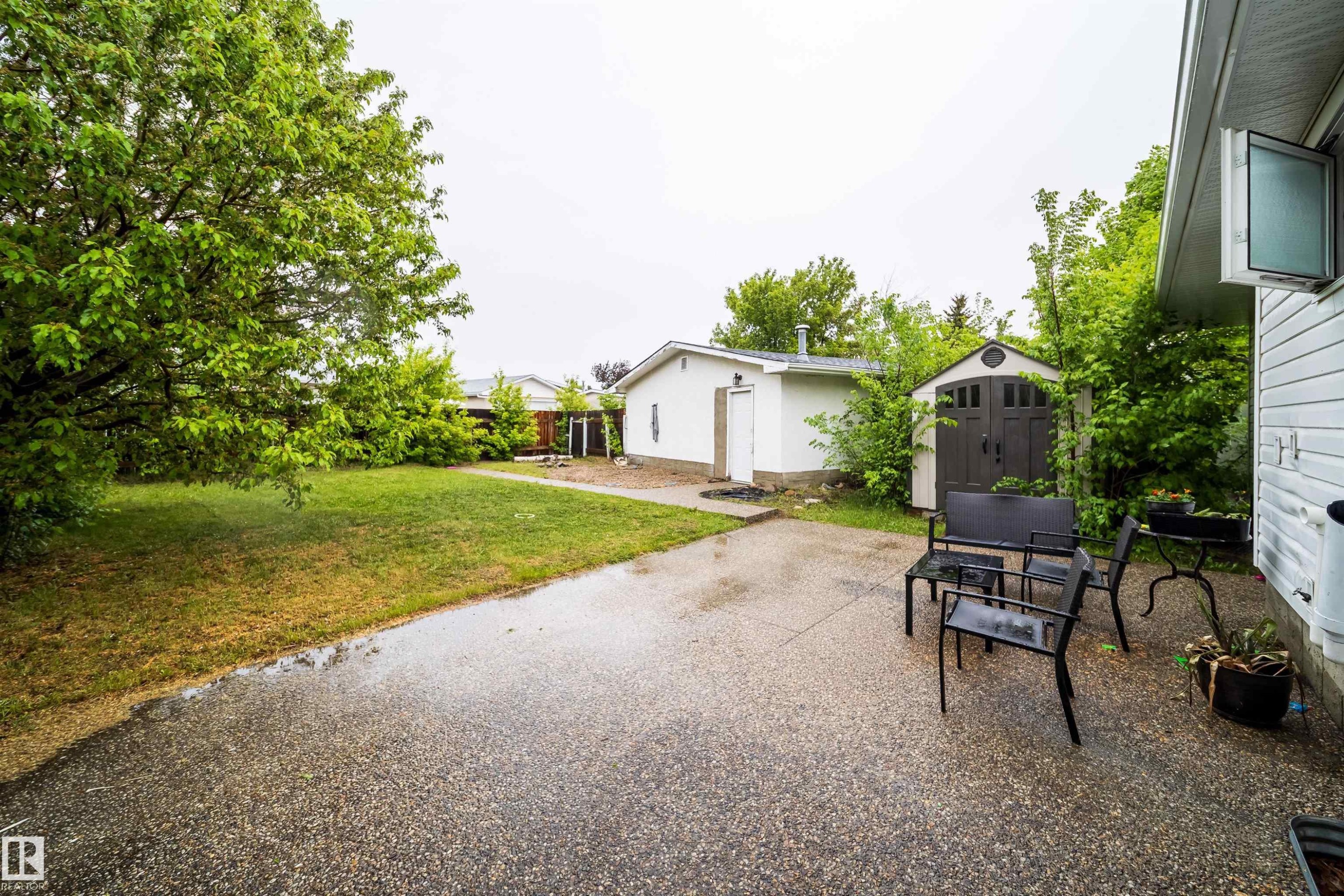 Expansive backyard featuring a large paved patio, a separate storage shed, a detached white auxiliary building, and mature perimeter trees - 8512 142A Avenue, Edmonton, AB