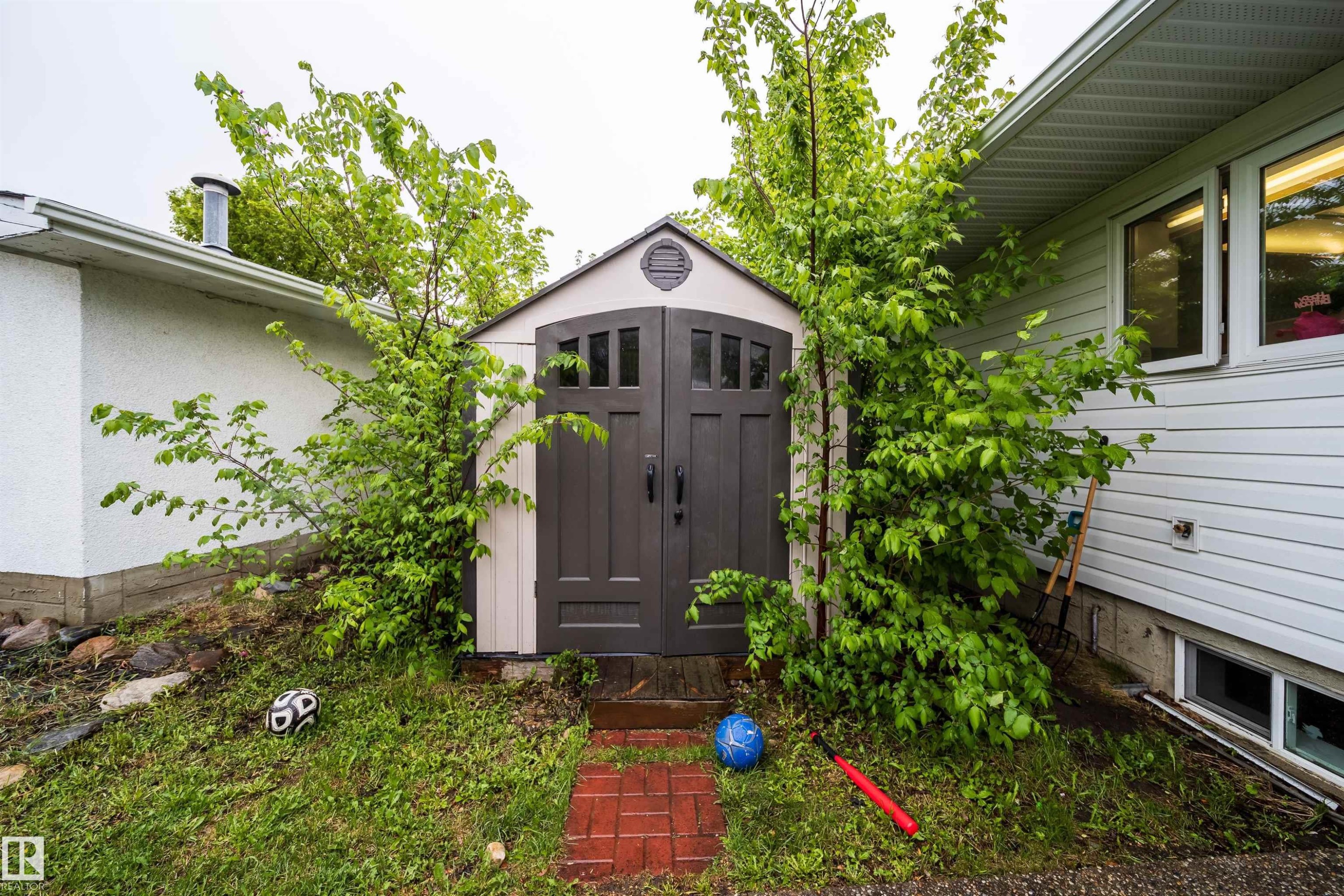 Freestanding shed with double arched doors and upper transom windows, set on a sloped grassy area with a brick pathway - 8512 142A Avenue, Edmonton, AB
