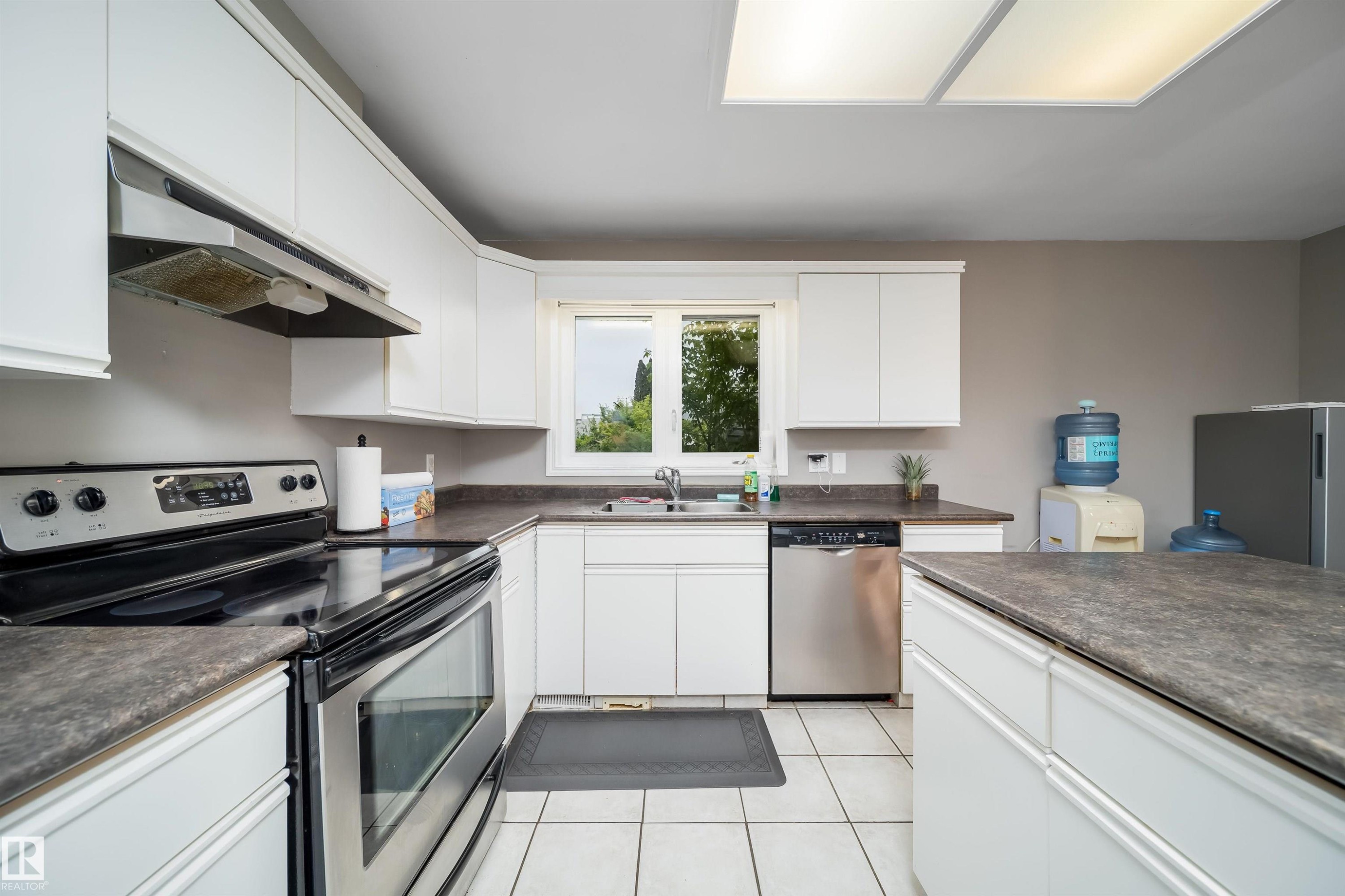 Kitchen featuring white cabinetry, dark countertops, white tile flooring, stainless steel appliances, and an island - 8512 142A Avenue, Edmonton, AB