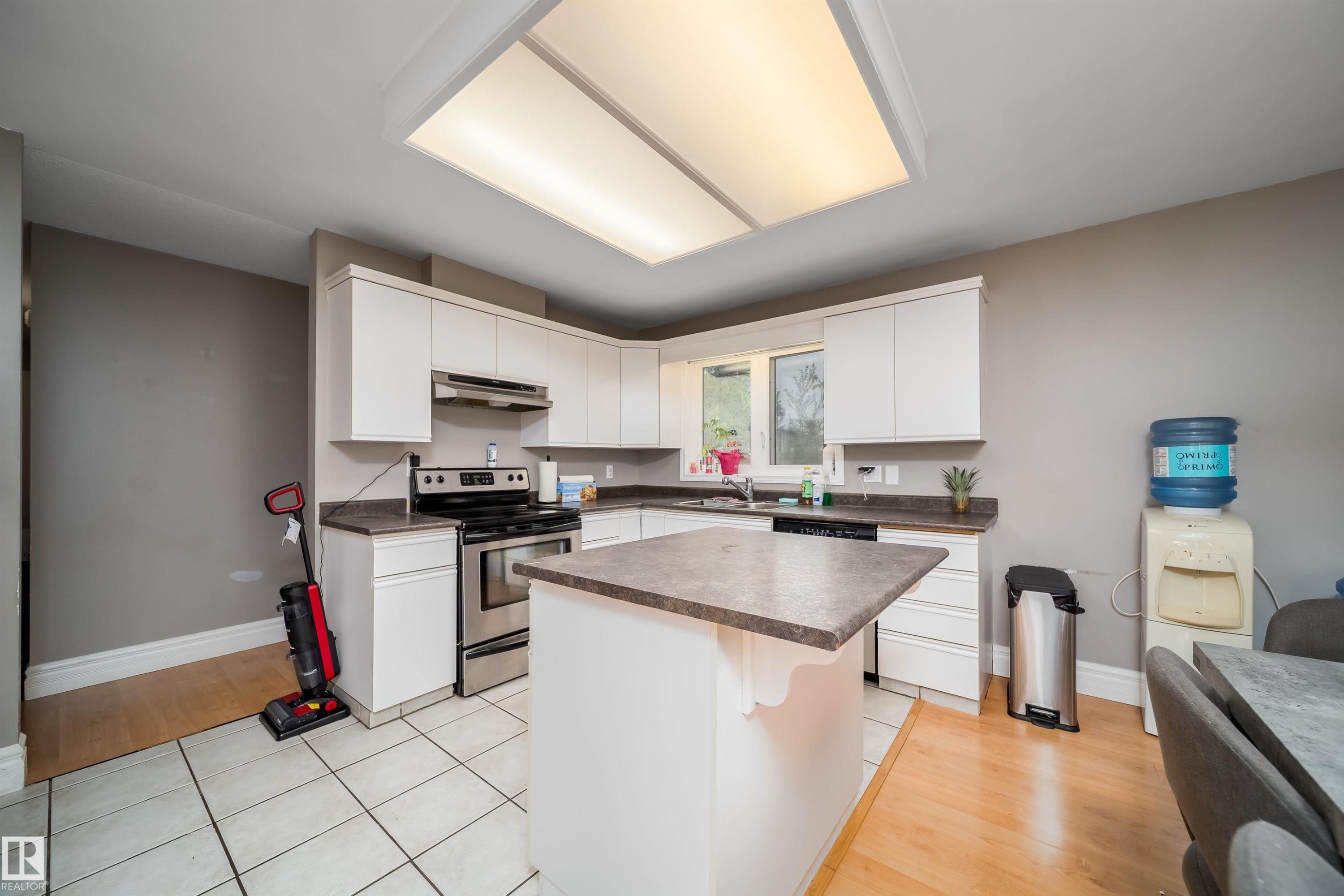 Kitchen featuring white cabinetry, stainless steel appliances, a center island with countertop, a window above the sink, and a combination of tile and wood-finish flooring - 8512 142A Avenue, Edmonton, AB