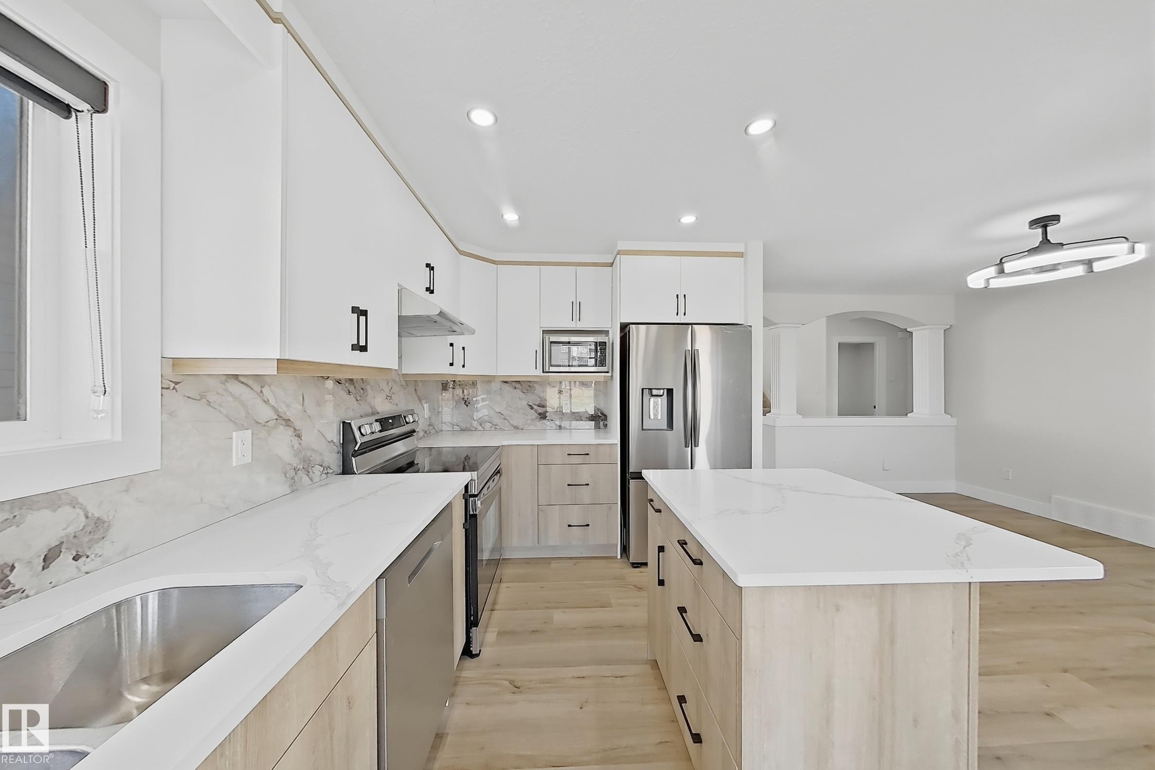 Kitchen featuring an oversized island with white countertop, stainless steel appliances, a white and wood-finish cabinetry scheme, and matching marble-patterned backsplash - 6144 166 Avenue, Edmonton, AB