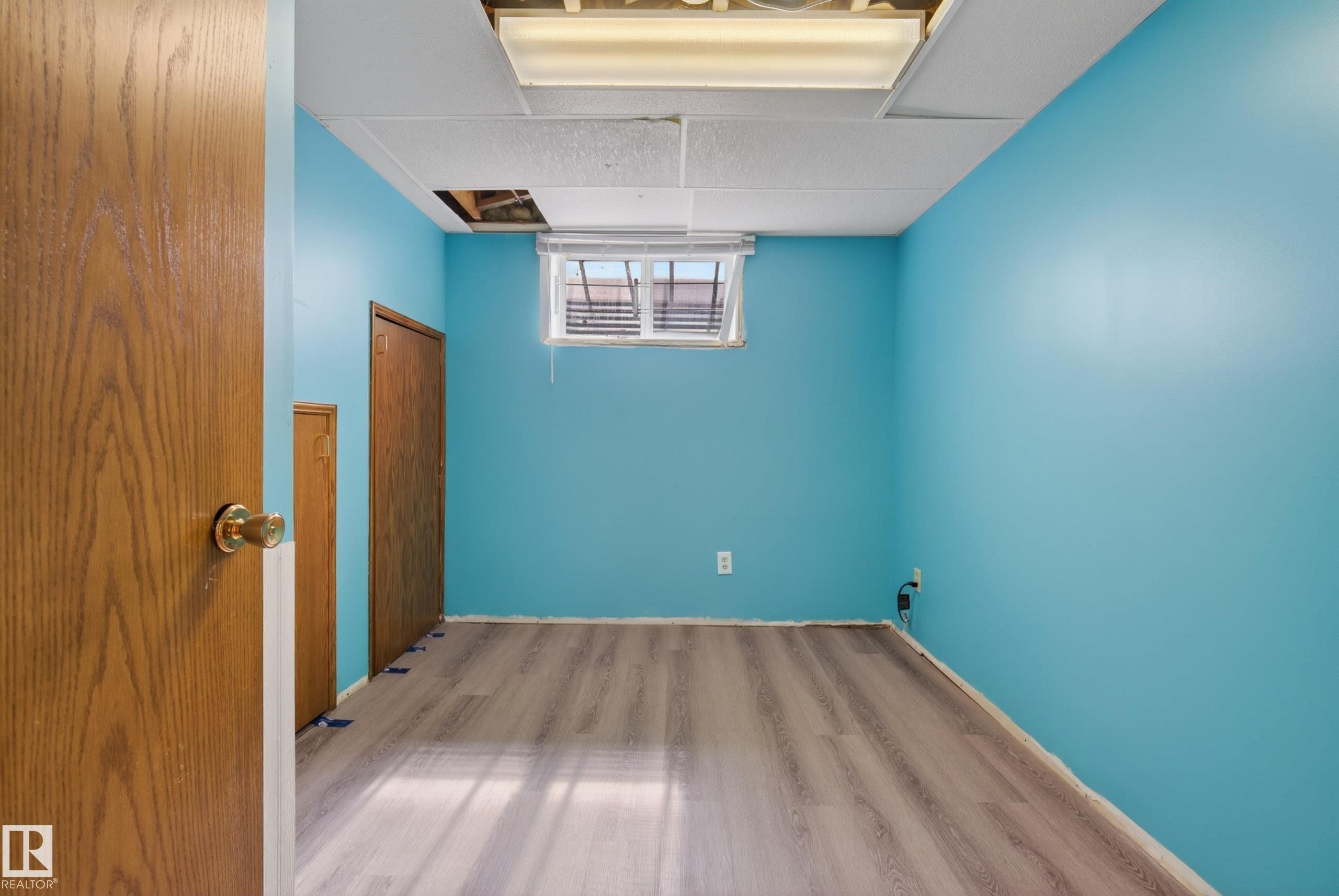Spacious room featuring wood-finish flooring, two wood-paneled doors, a window with horizontal blinds, and a drop ceiling with integrated fluorescent lighting - 6203 90 Avenue, Edmonton, AB