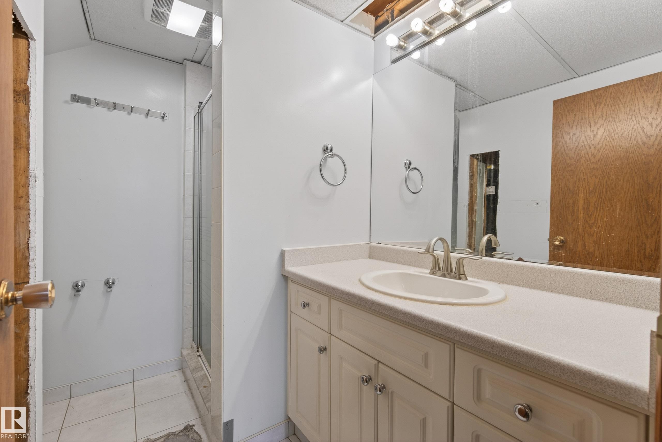 Bathroom featuring an integrated vanity sink with a brushed nickel faucet, a spacious mirror with overhead bar lighting, a shower enclosure with a glass door, and a tiled floor - 6203 90 Avenue, Edmonton, AB