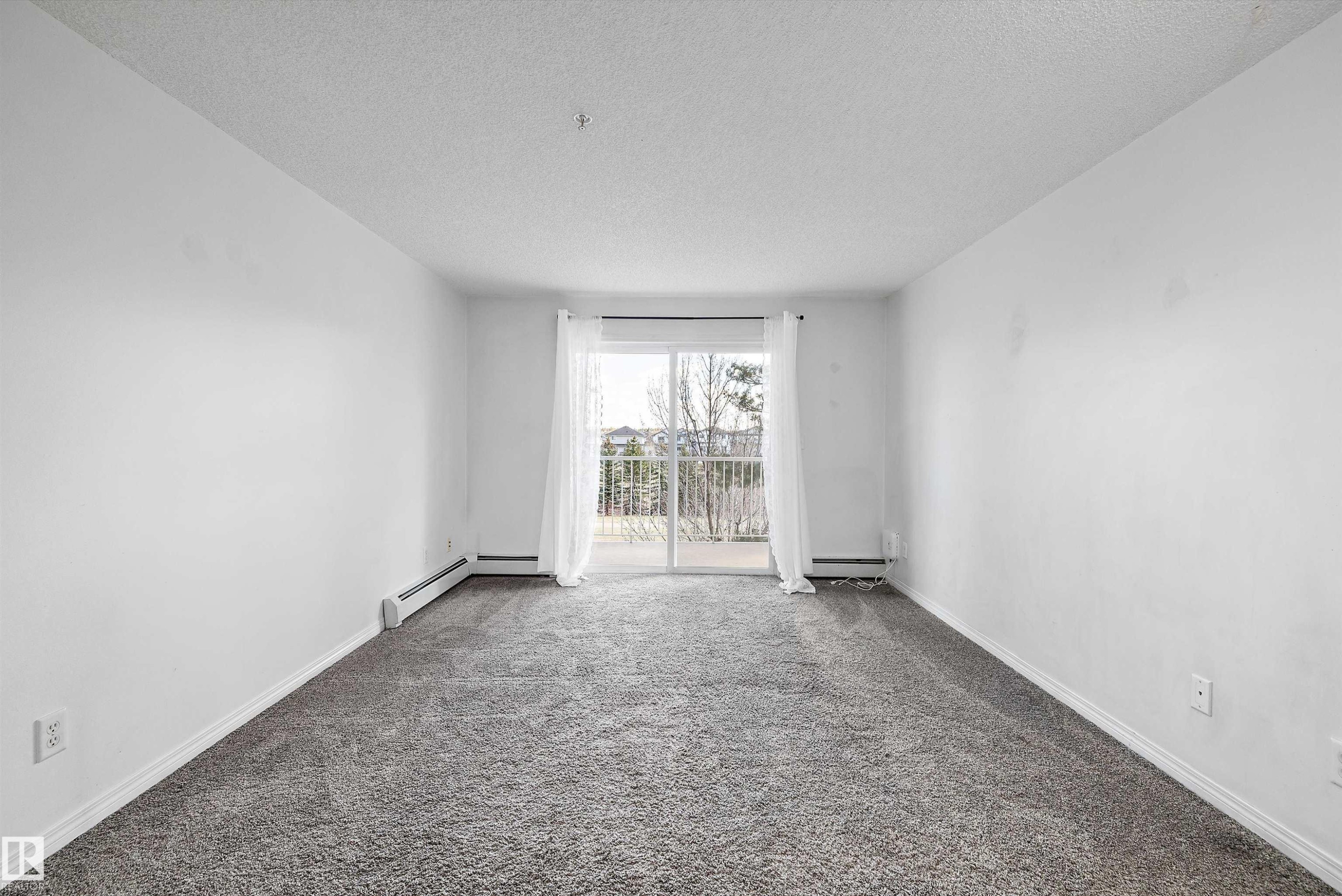 Spacious room featuring gray textured carpet, white painted walls, and a sliding glass door leading to an outdoor area with a white railing - 320 245 Edwards Drive, Edmonton, AB