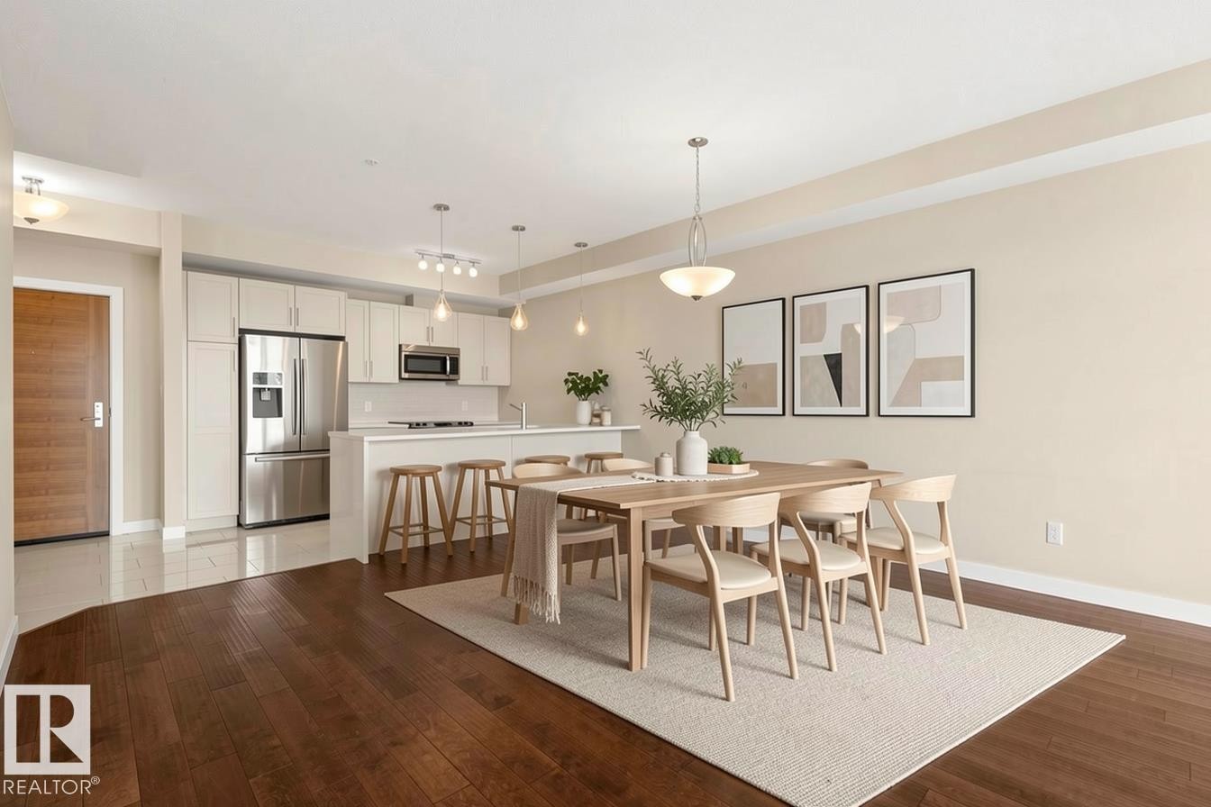 Open-concept living space featuring wood-finish flooring, a contemporary kitchen with stainless steel appliances, white cabinetry, and a central island with bar seating - 309 625 Leger Way, Edmonton, AB