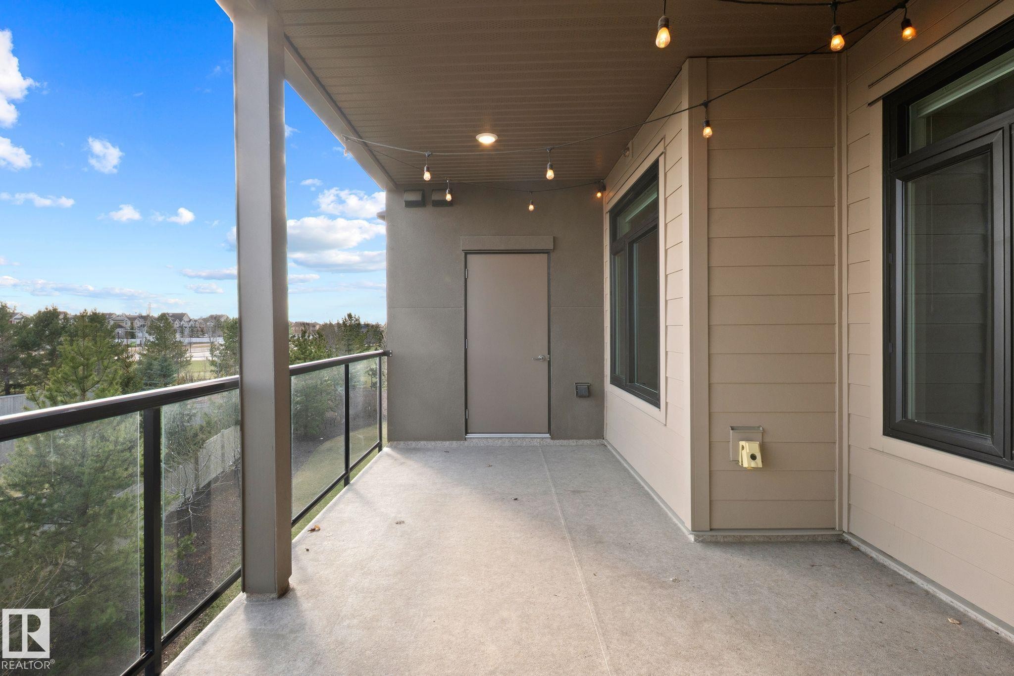 Spacious exterior balcony featuring a concrete surface, glass panel railing, overhead string lighting, and direct outdoor access - 309 625 Leger Way, Edmonton, AB