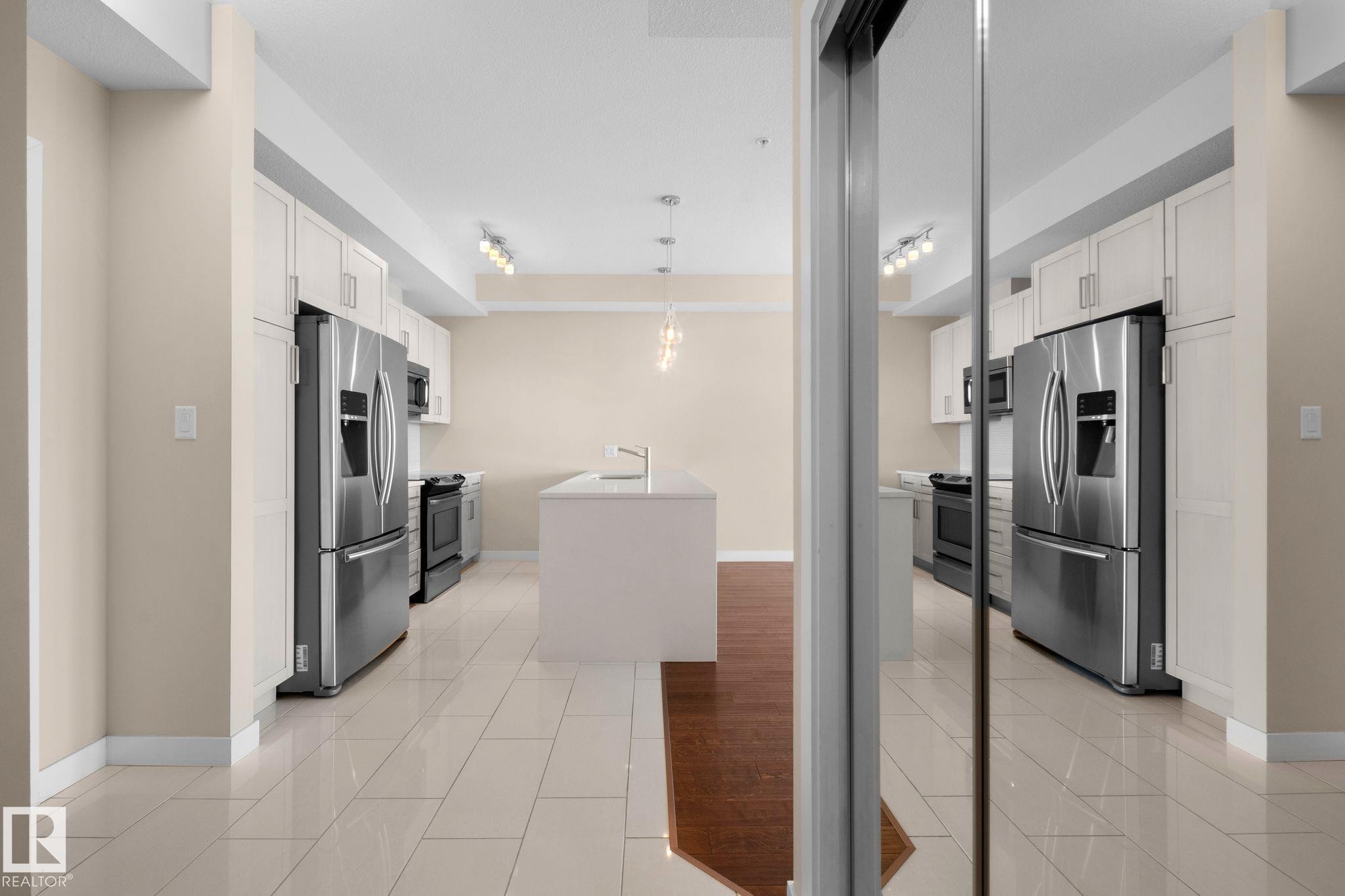 Modern kitchen featuring an expansive center island, stainless steel appliances, white cabinetry, track lighting, and light-toned tile flooring - 309 625 Leger Way, Edmonton, AB