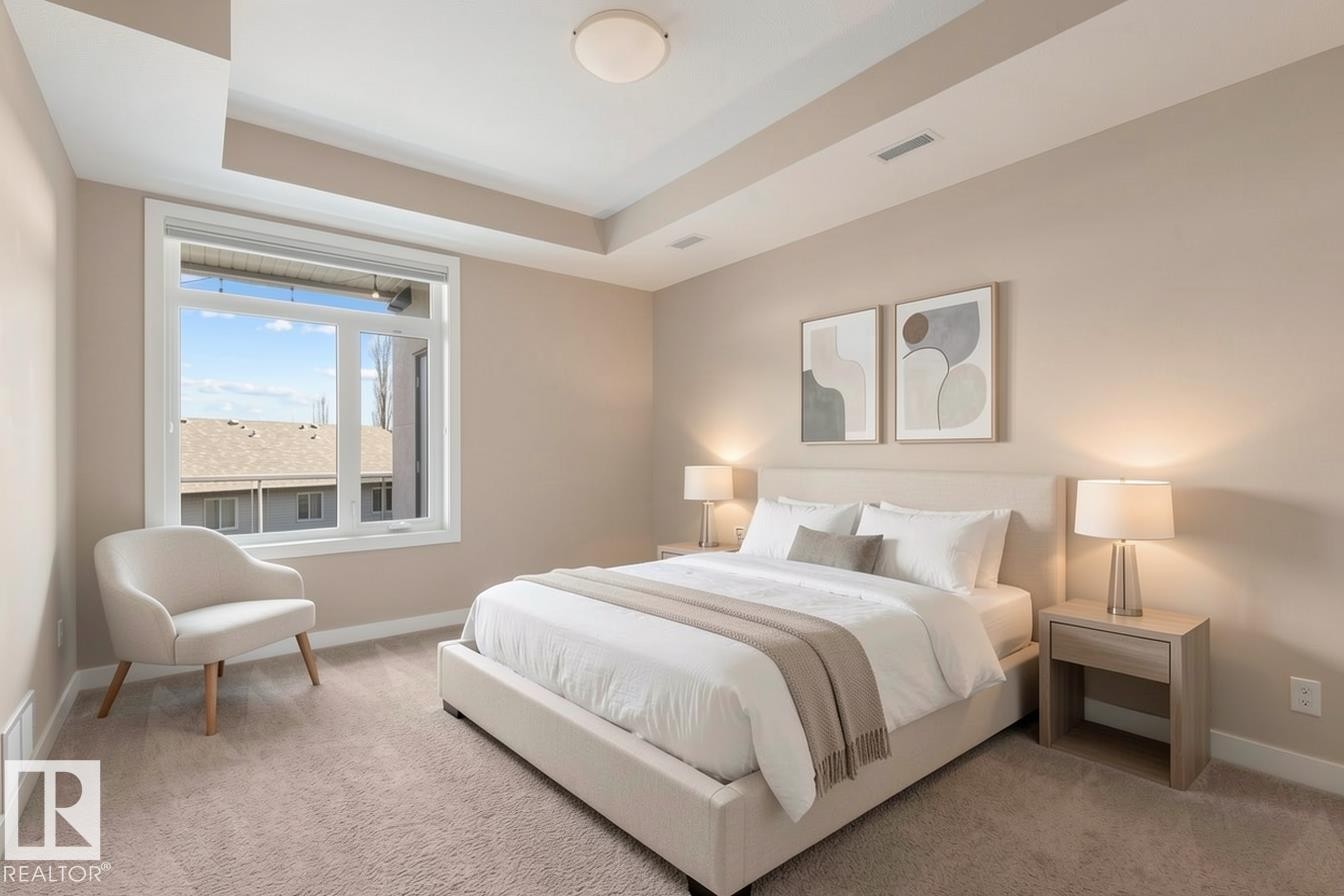 Spacious bedroom featuring a large window, tray ceiling, neutral-toned walls, recessed lighting, and soft carpet flooring - 309 625 Leger Way, Edmonton, AB