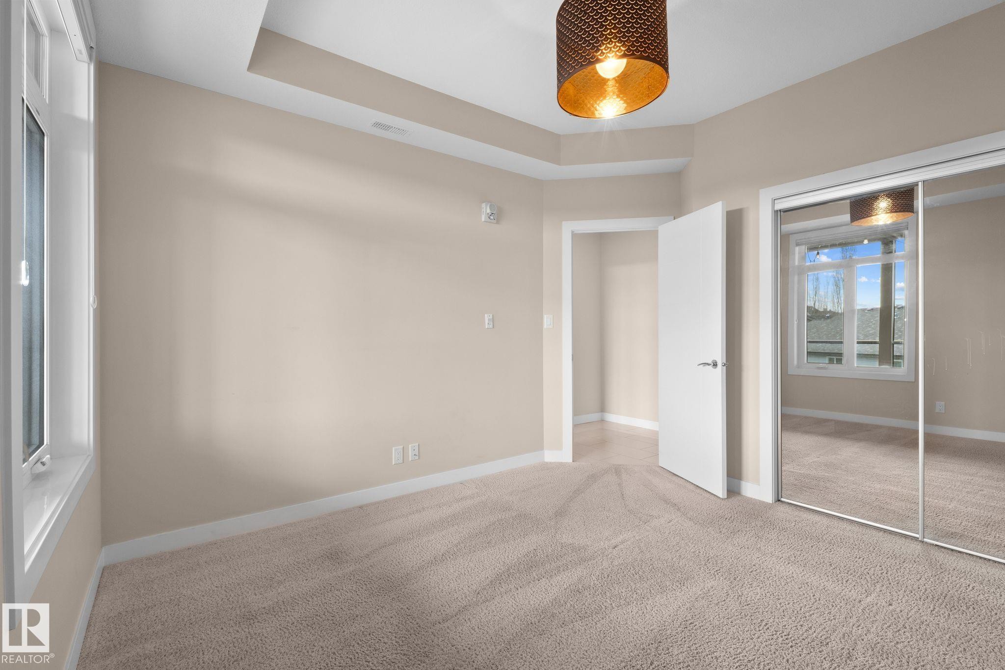 Carpeted room featuring a tray ceiling, a drum pendant light fixture, a window with white trim, and mirrored sliding closet doors - 309 625 Leger Way, Edmonton, AB